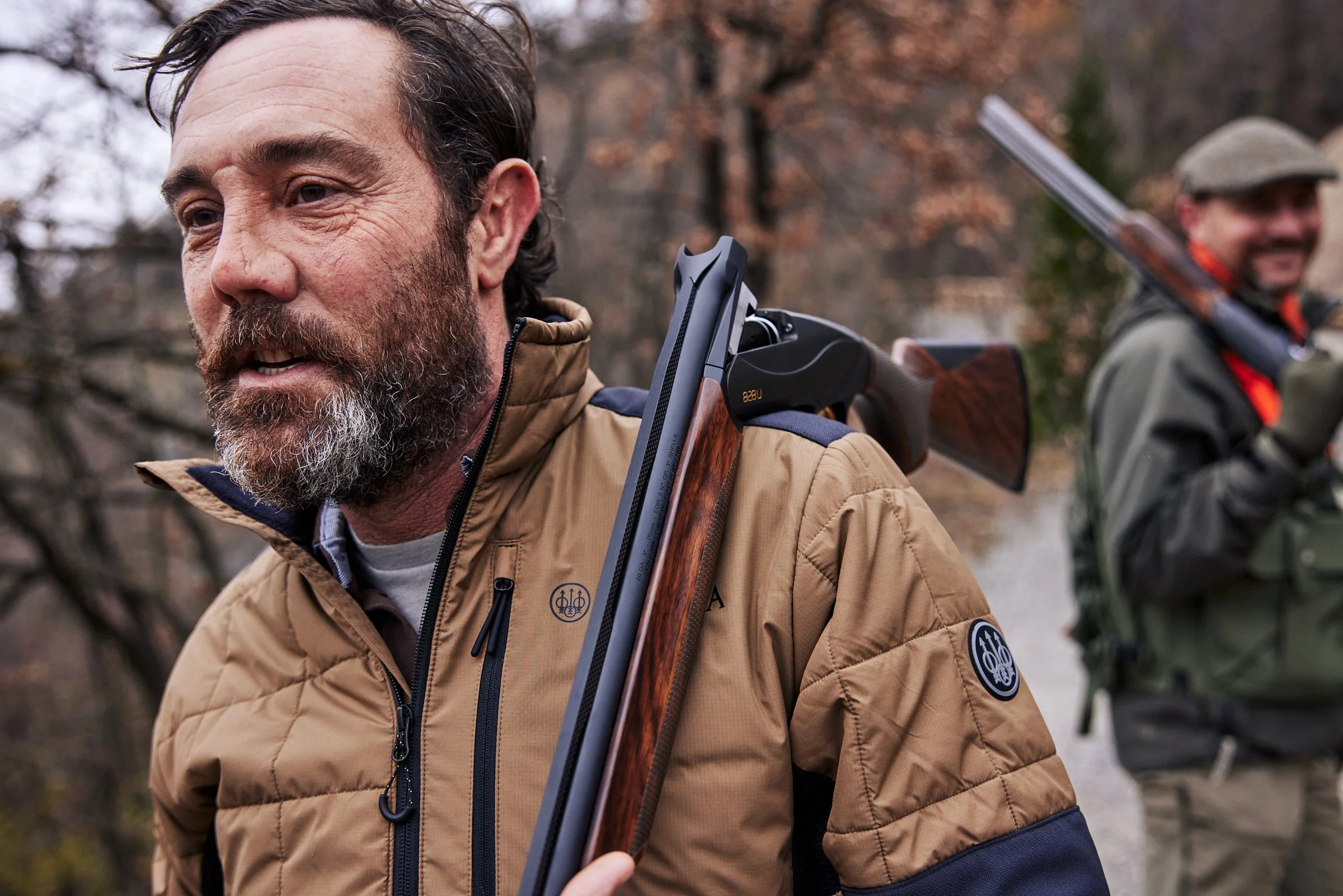 Two men outdoors with rifles on their shoulders, dressed in outdoor jackets, in a forested area.