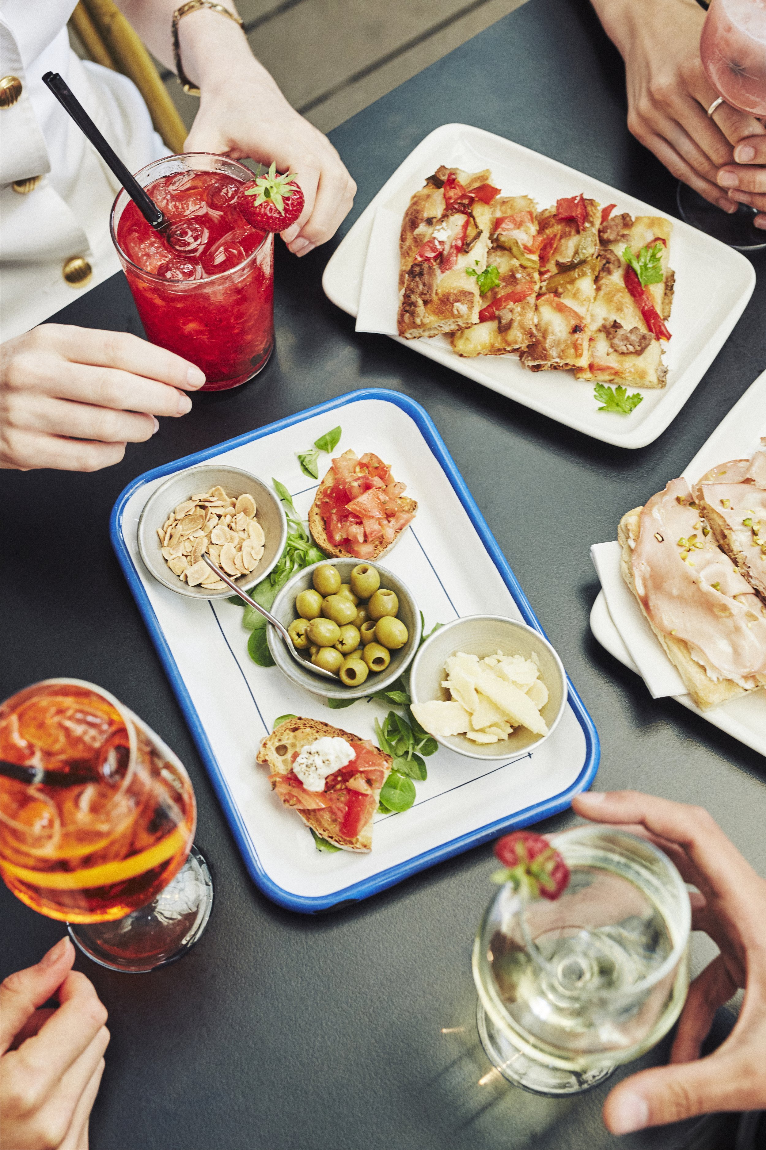 A group of people enjoying a meal at an outdoor table. The table is set with various dishes, including flatbread with toppings, bruschetta with cherry tomatoes and cheese, bowls of olives, nuts, and cheese slices. There are also assorted drinks inclu