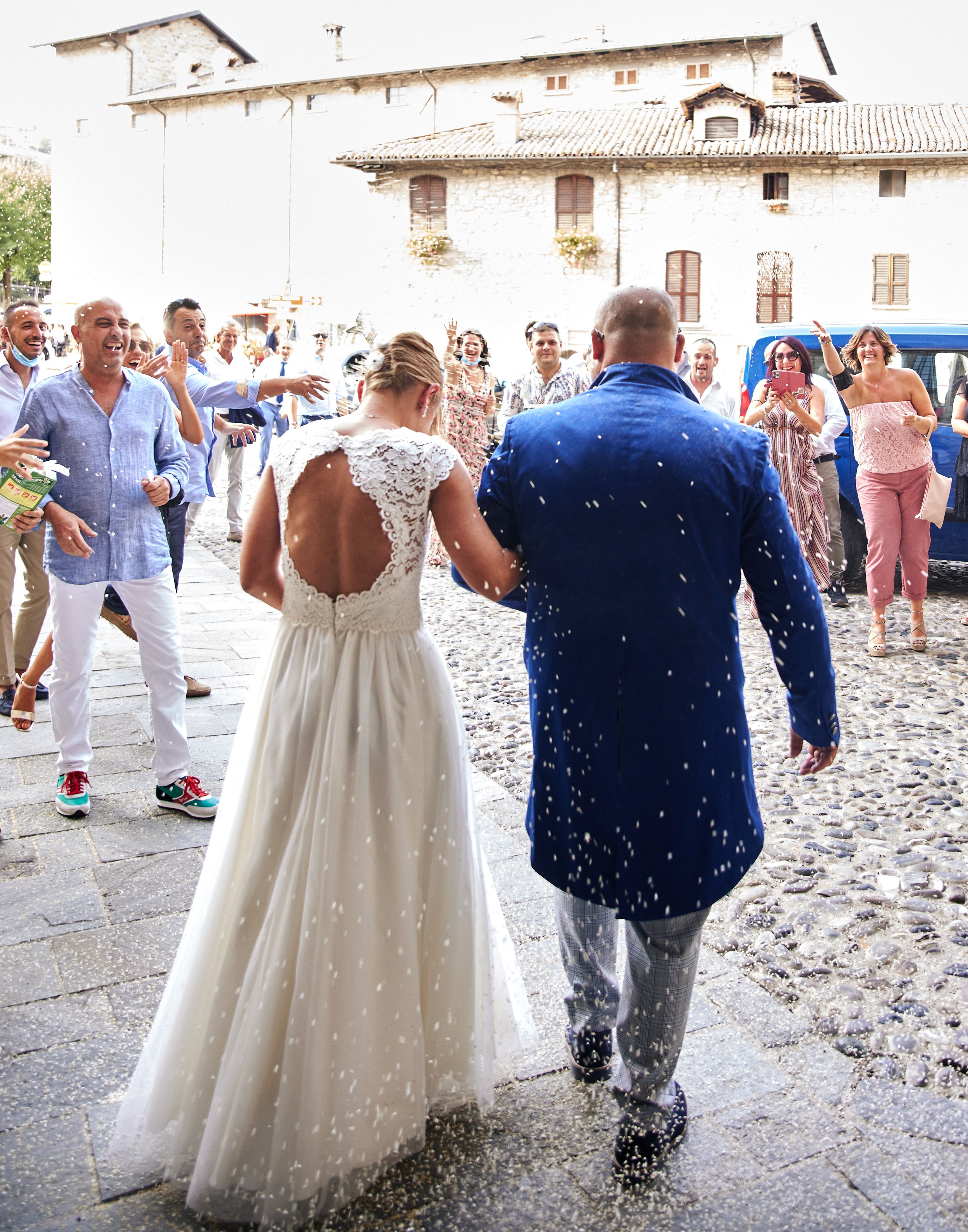 Bride and groom walking away, guests throwing rice, cobblestone street, festive atmosphere.