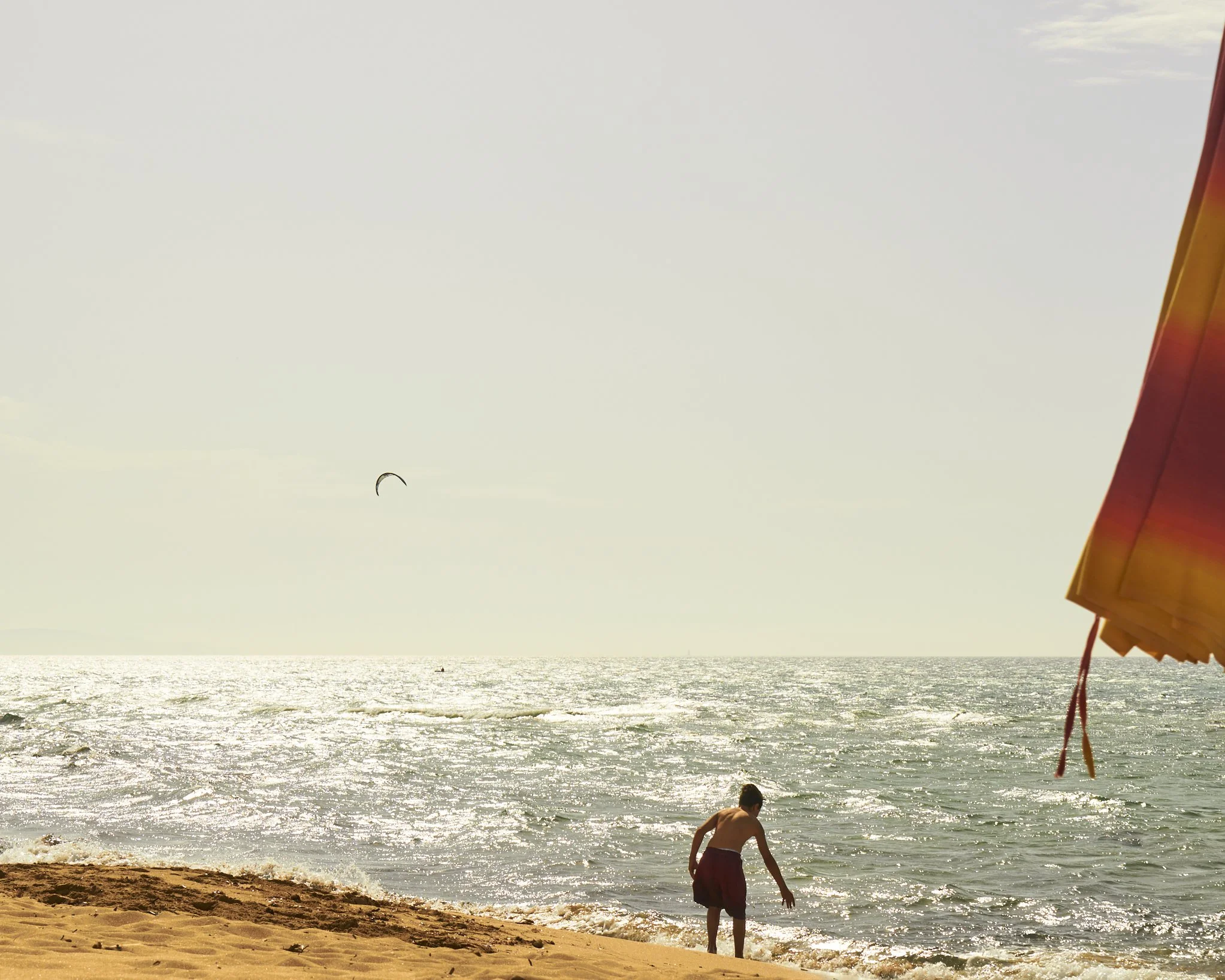 A person playing by the ocean on a sandy beach under a clear sky with a kite in the background and a striped beach umbrella on the side.