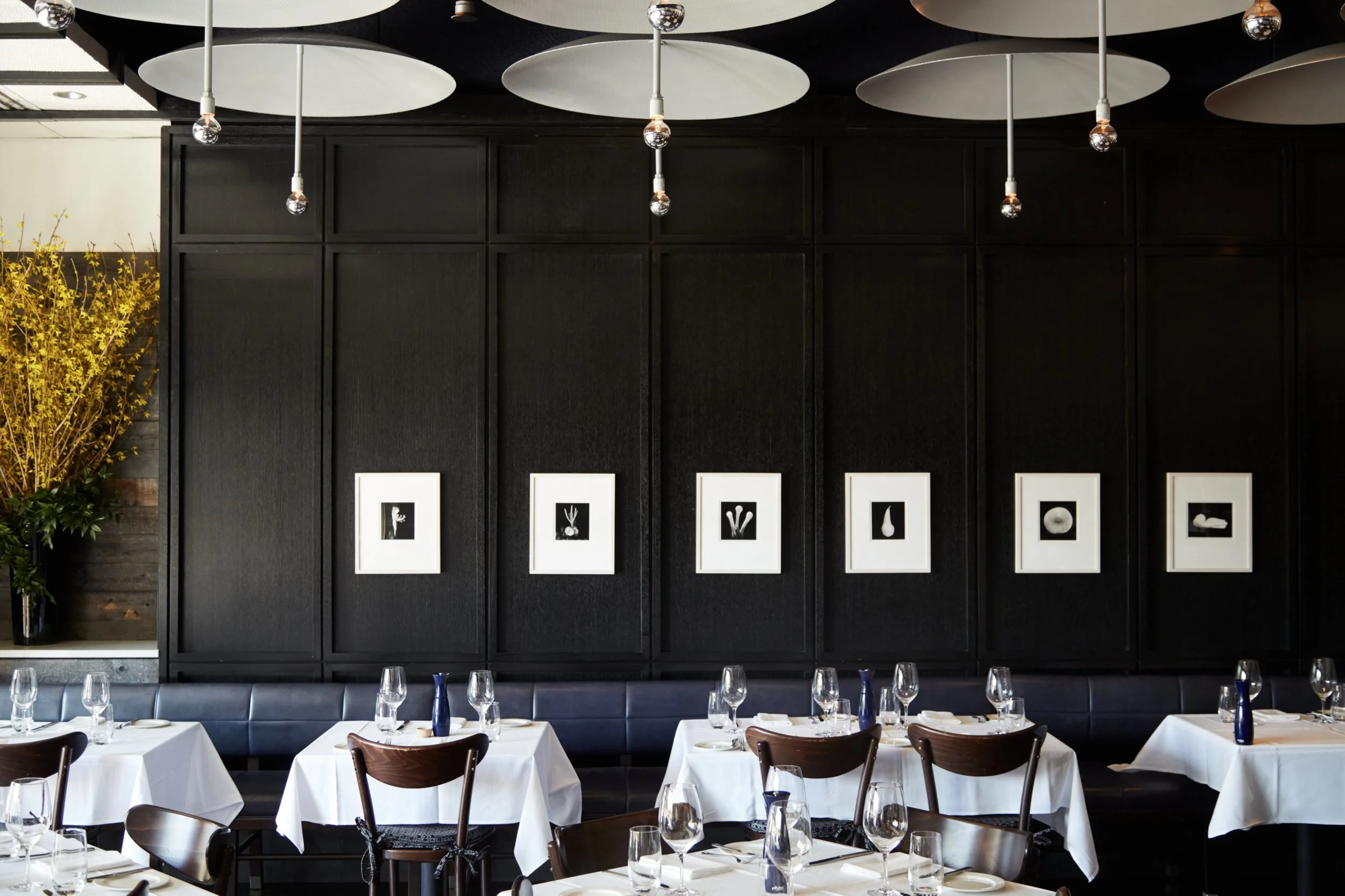Elegant restaurant interior with dark walls, white tablecloths, and monochrome artwork. Ceiling features round light fixtures. Tables are set with wine glasses and silverware.