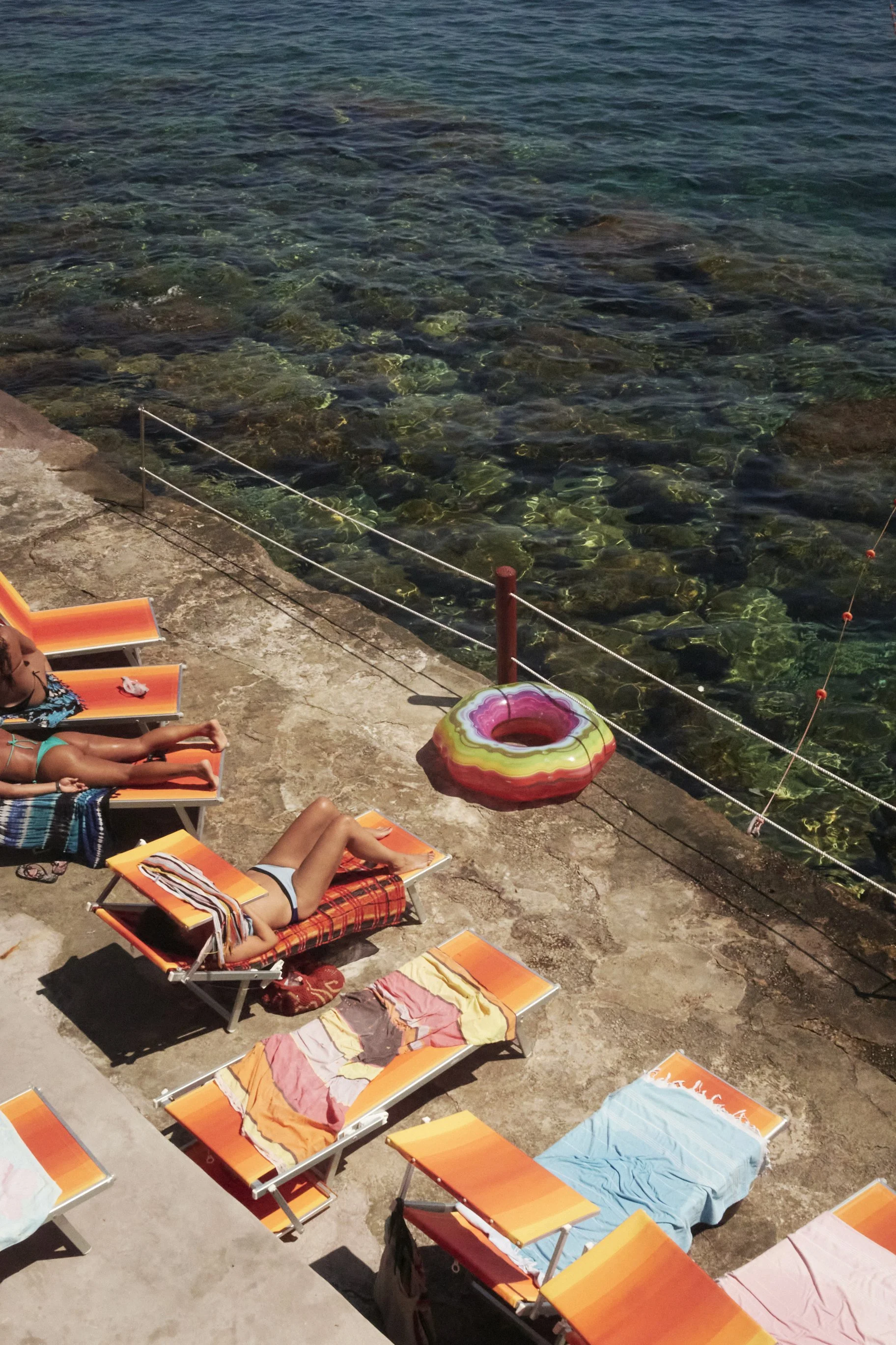People sunbathing on orange lounge chairs by the sea with a colorful inflatable ring nearby.