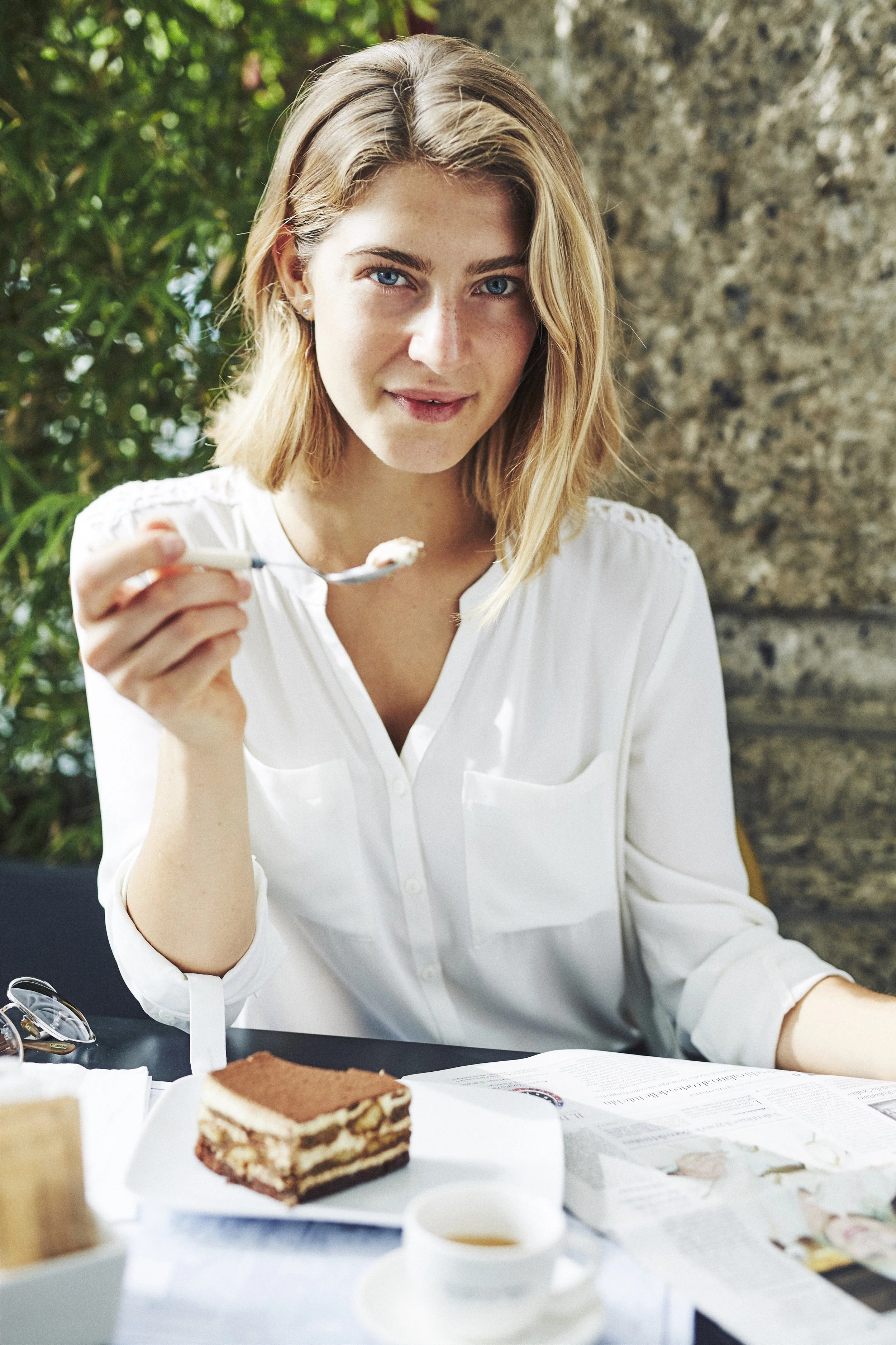 Woman wearing a white blouse eating dessert at a table with tiramisu, coffee, and a newspaper.