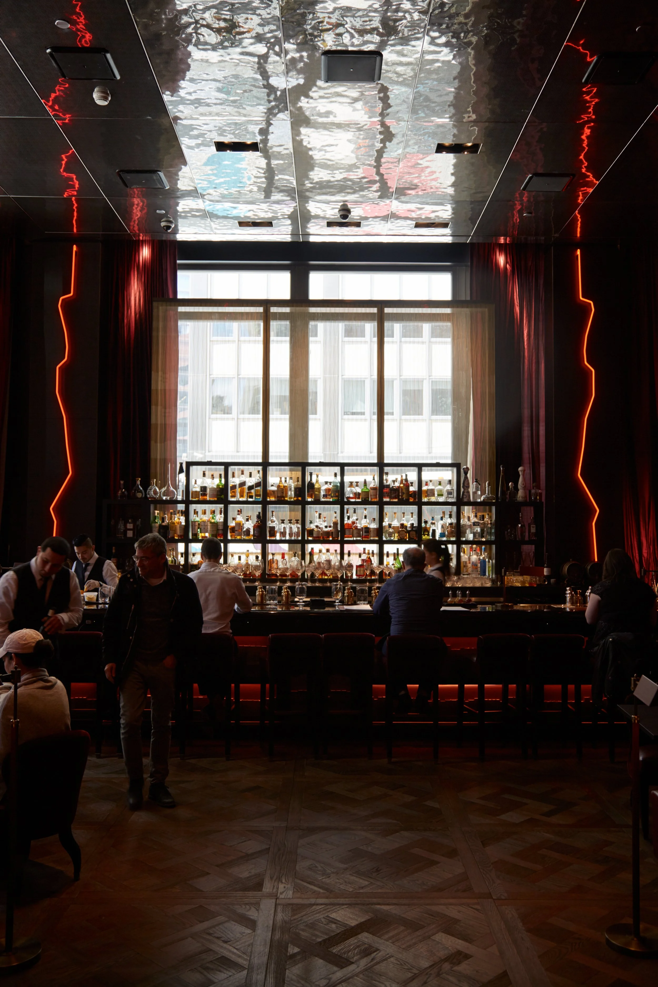 Stylish bar interior with patrons seated at the counter, rows of liquor bottles on shelves, and a reflective ceiling with red neon lighting accents.