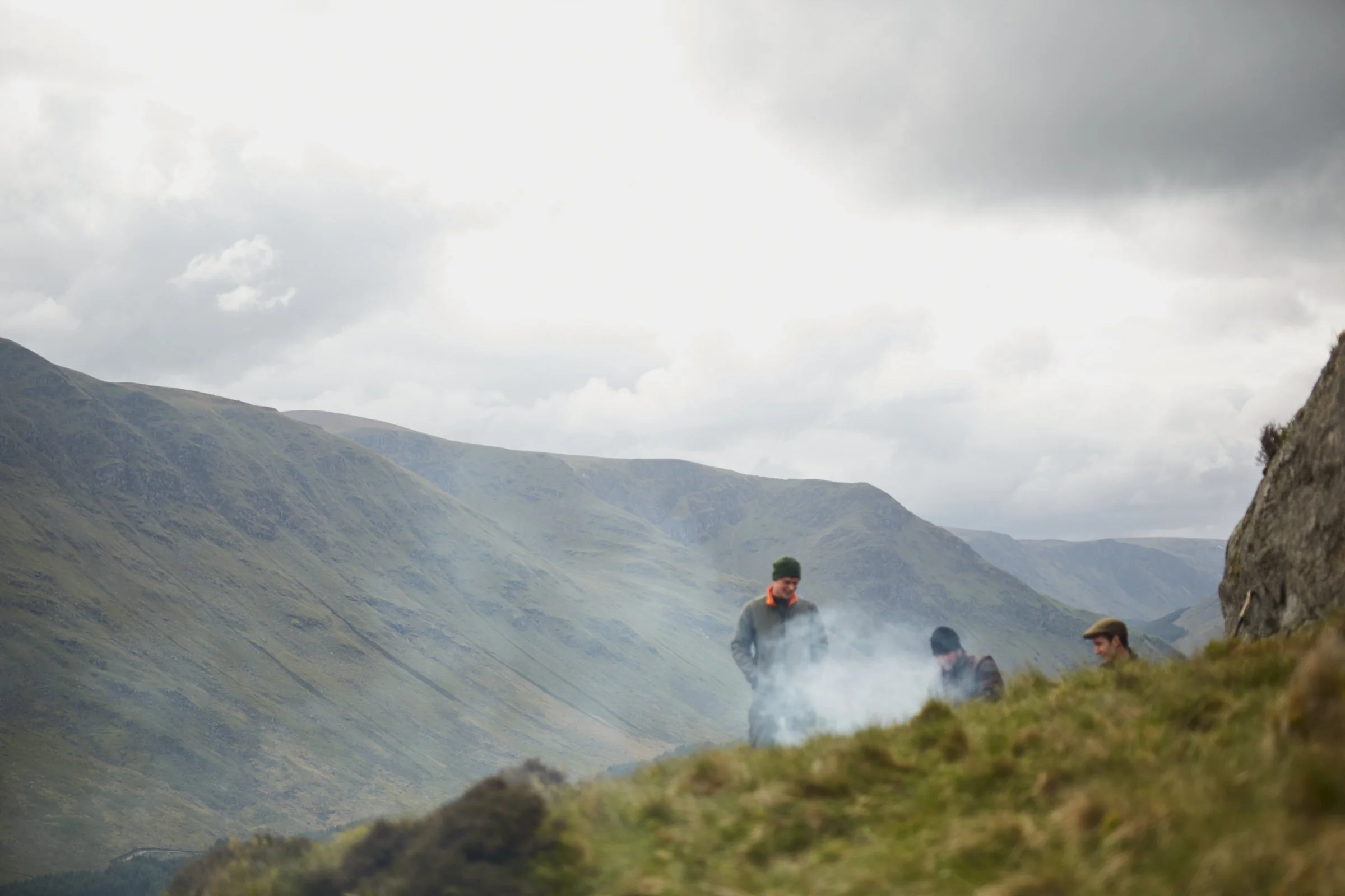 Three people on a grassy hill with smoke, overlooking a mountainous landscape under a cloudy sky.