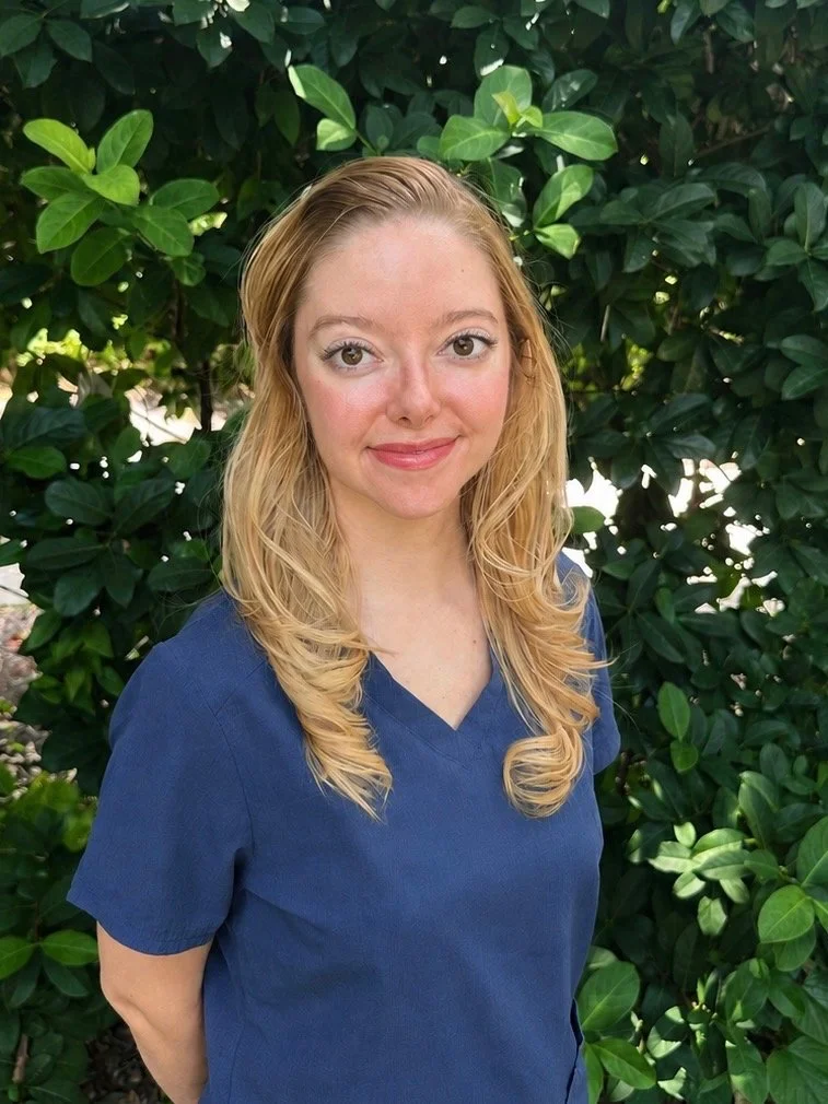 A young woman with long, wavy blonde hair standing outdoors in front of green leafy bushes, wearing a blue top.