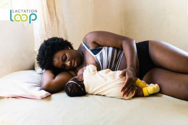 A mother reclines on a bed, gazing lovingly at her newborn baby while breastfeeding.