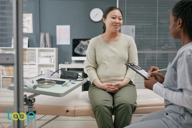 A pregnant woman sits on an exam table during a prenatal consultation, speaking with a lactation consultant who is taking notes.