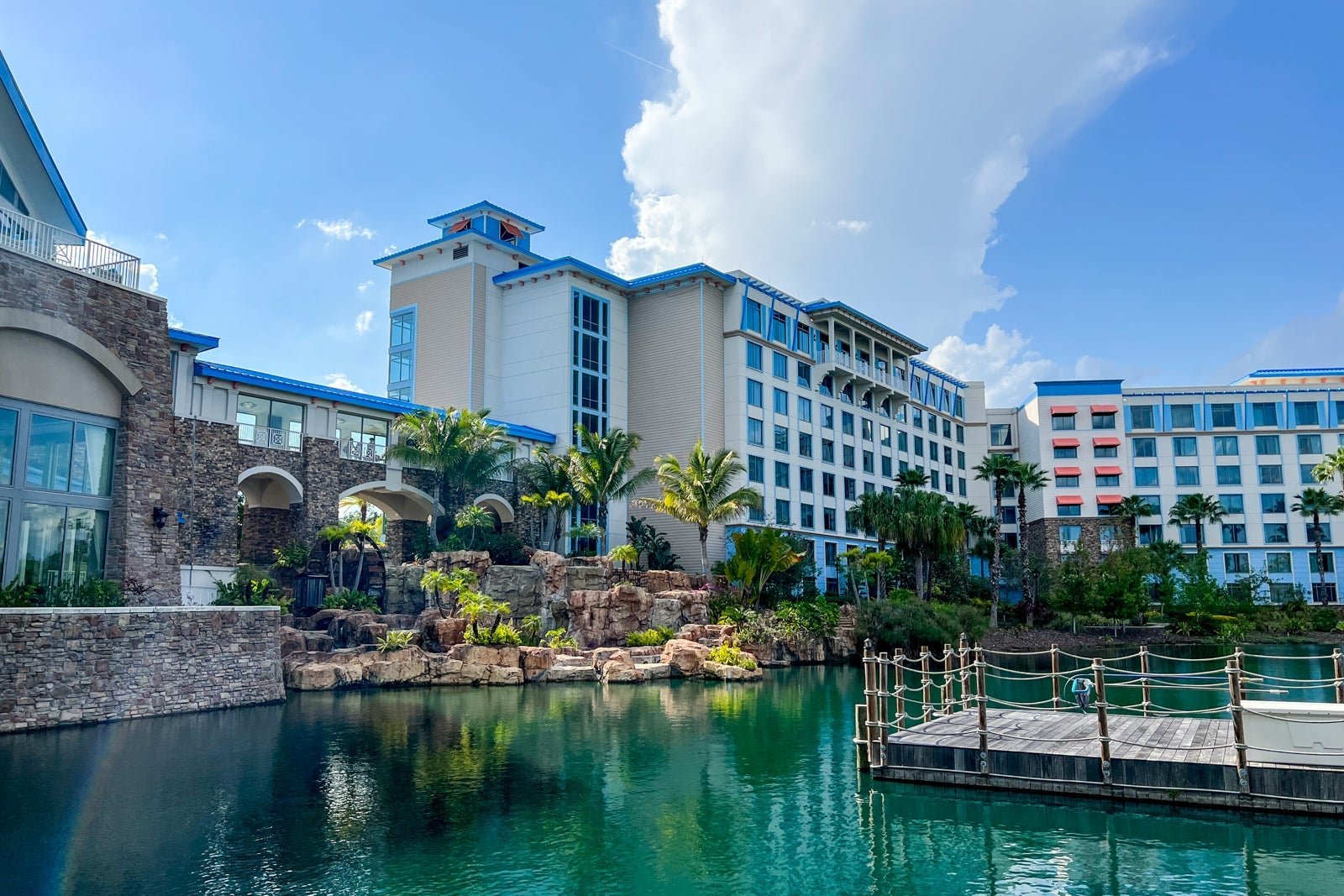 Luxury hotel with white and beige facade, blue accents, surrounded by palm trees, with a pond and dock in the foreground under a partly cloudy sky.