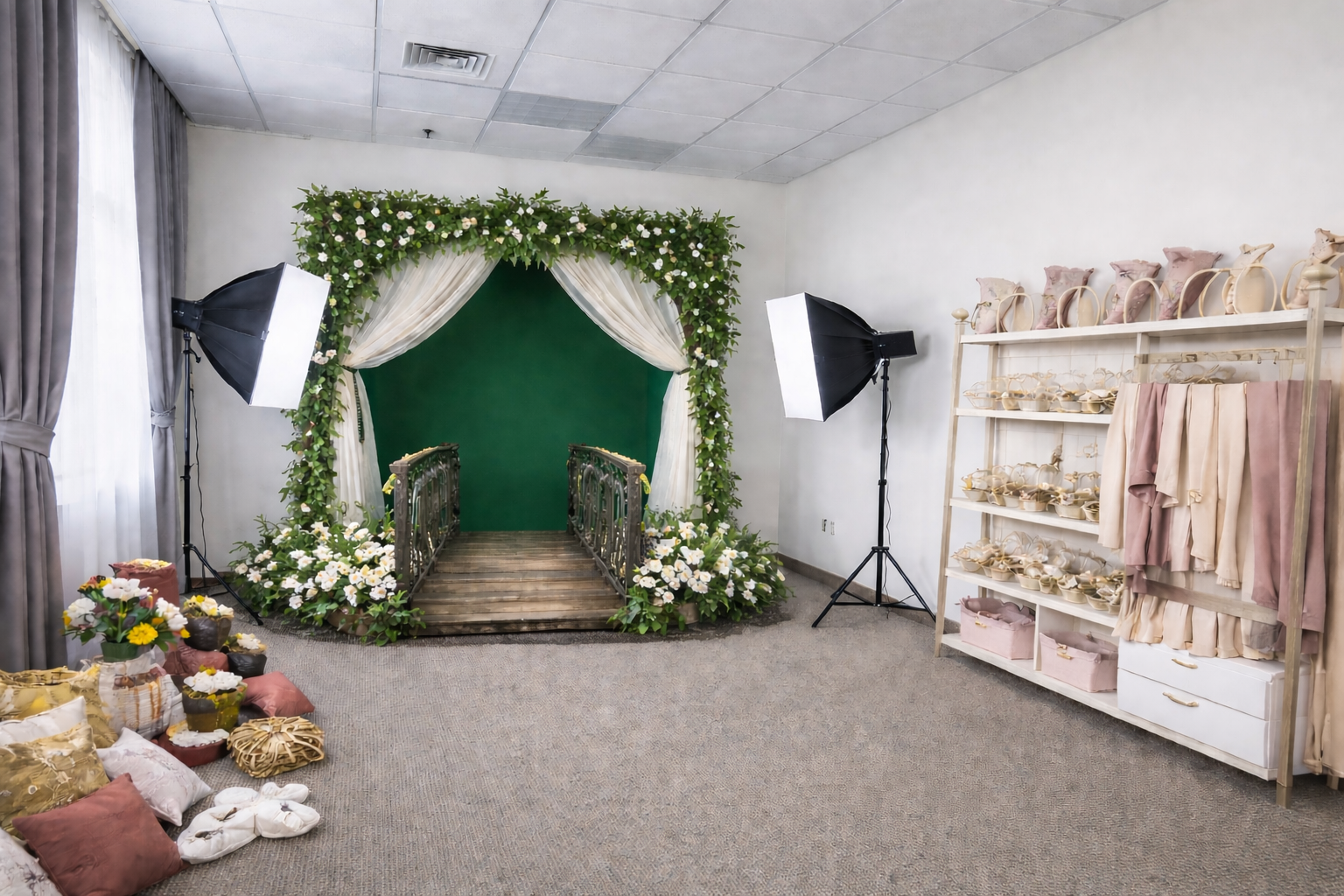 Indoor photography setup with a small decorative stage featuring floral arrangements, a miniature bridge, green backdrop, studios flashes, and props