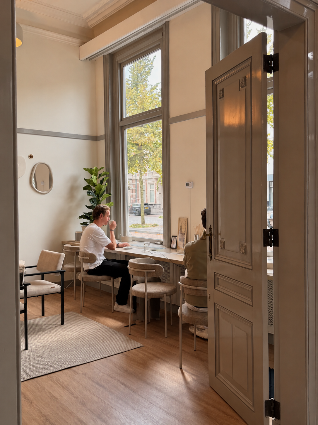 Interieur van een lichte, moderne koffieshop met grote ramen, planten, een leren bank, marmeren tafels, en stoelen. Ruimte bij Ivy Office.
