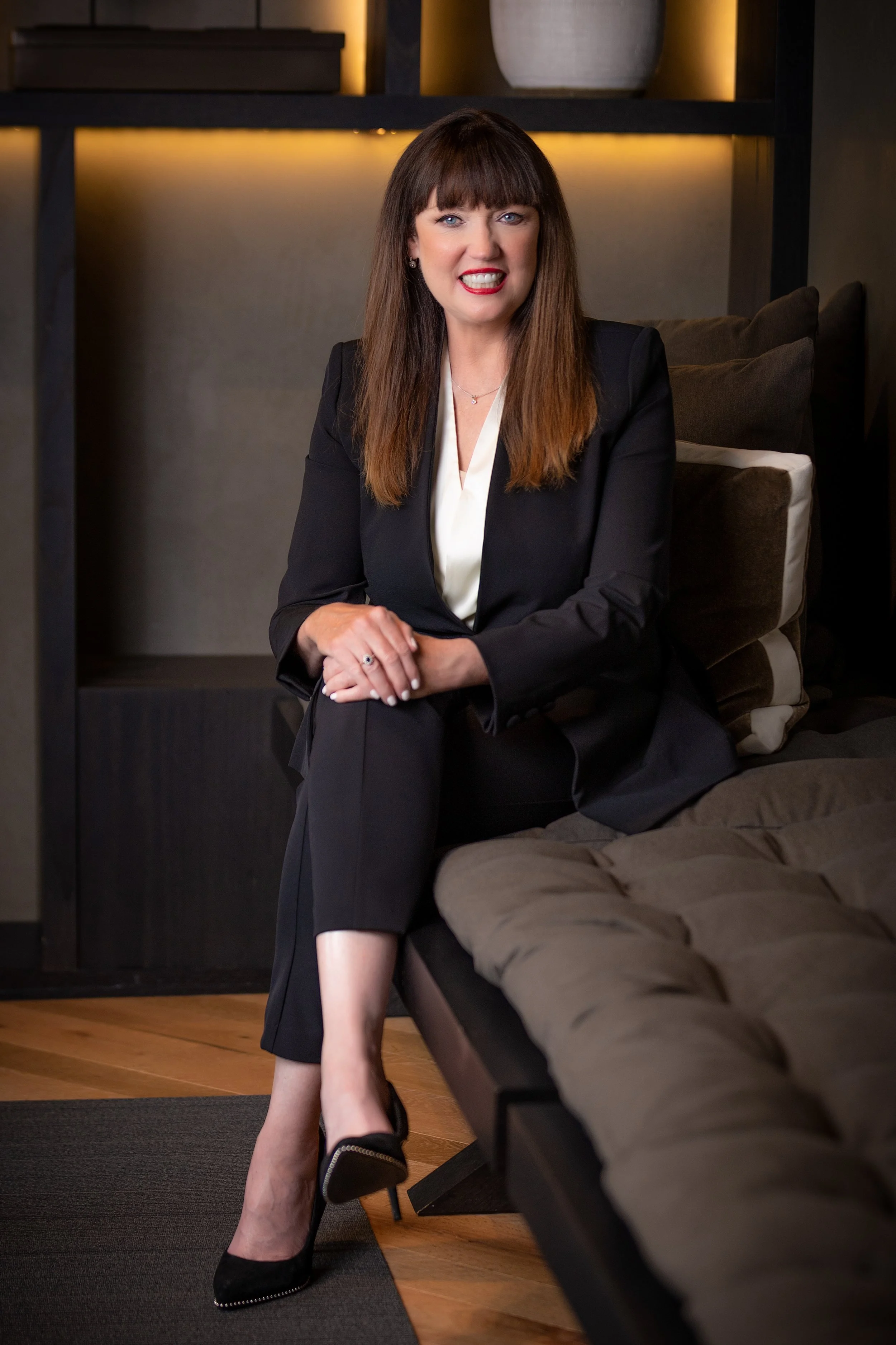 Woman in a black suit sitting on a couch in a modern interior with warm lighting.