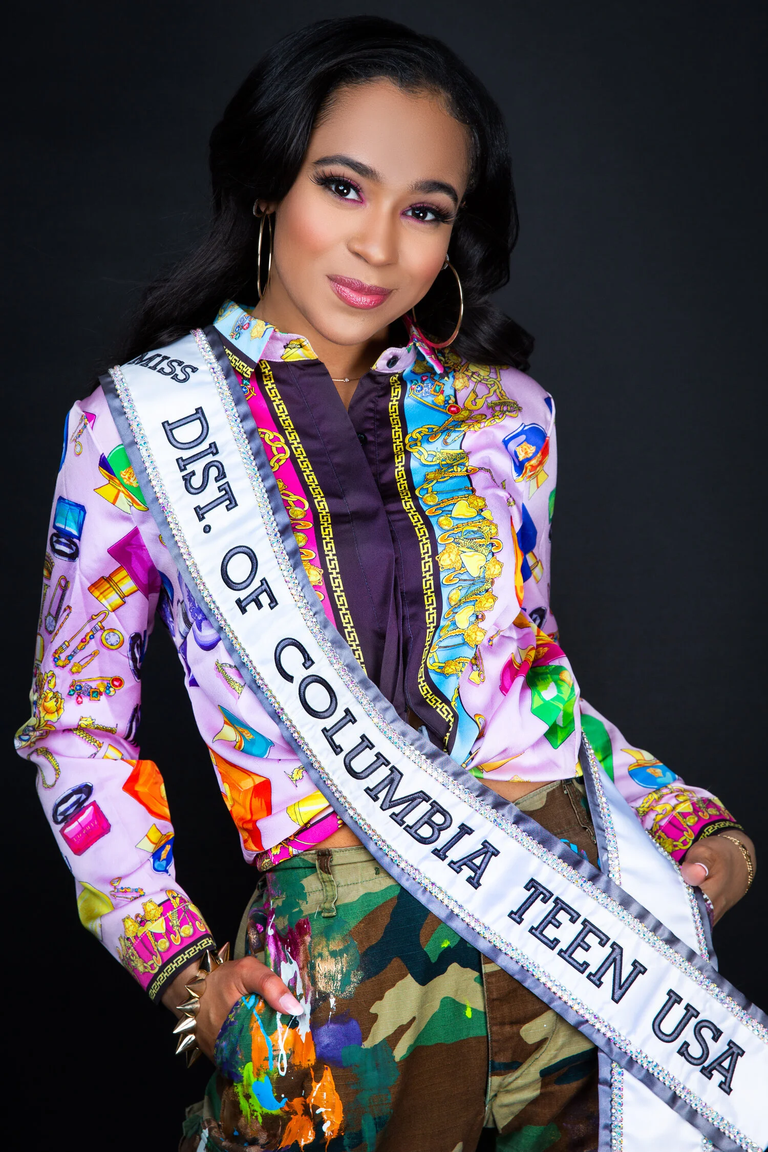 Person wearing a colorful patterned shirt and camo pants with a sash that reads 'Miss Dist. of Columbia Teen USA.'