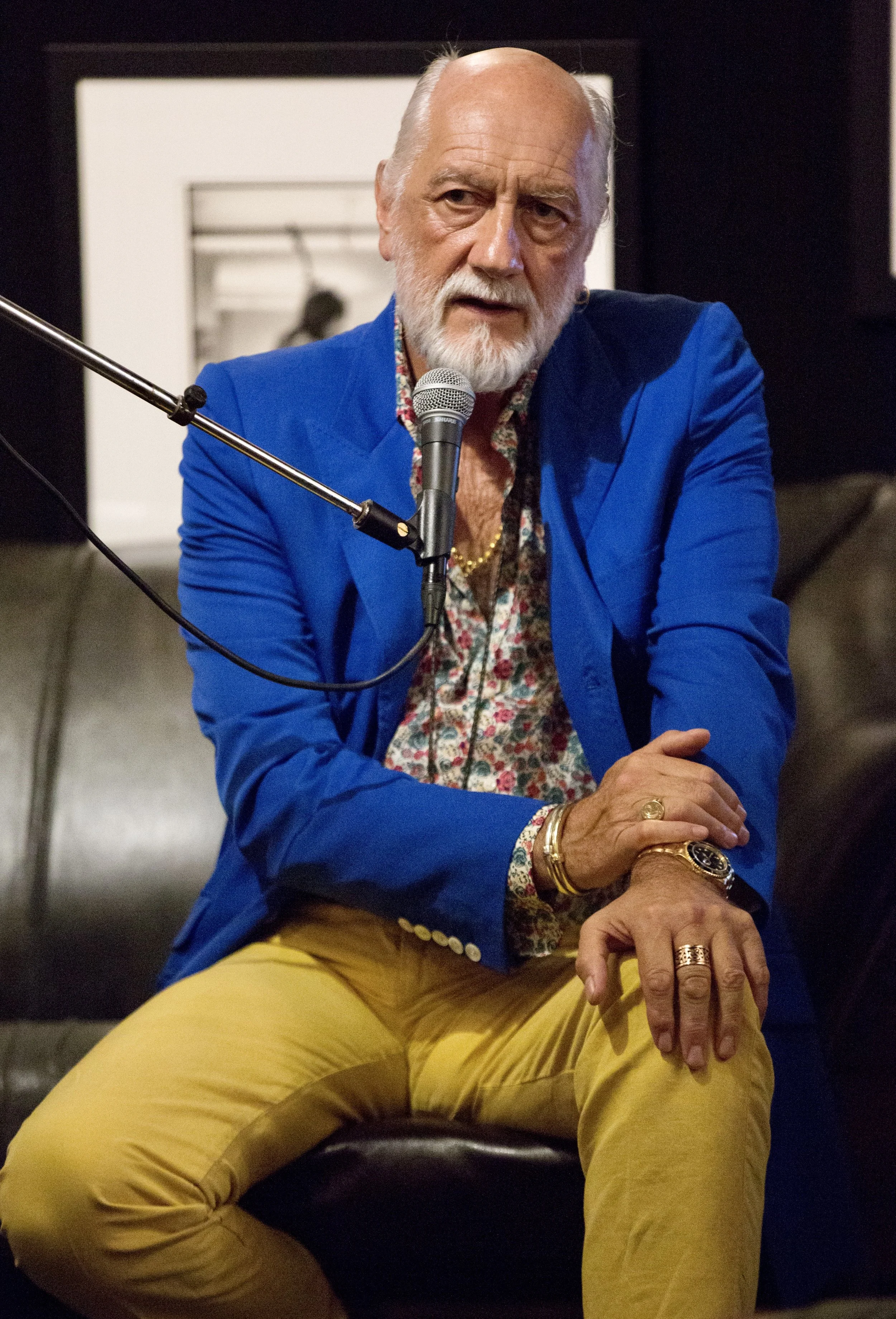 Older man with a white beard seated, wearing a blue blazer, floral shirt, and yellow pants, speaking into a microphone.