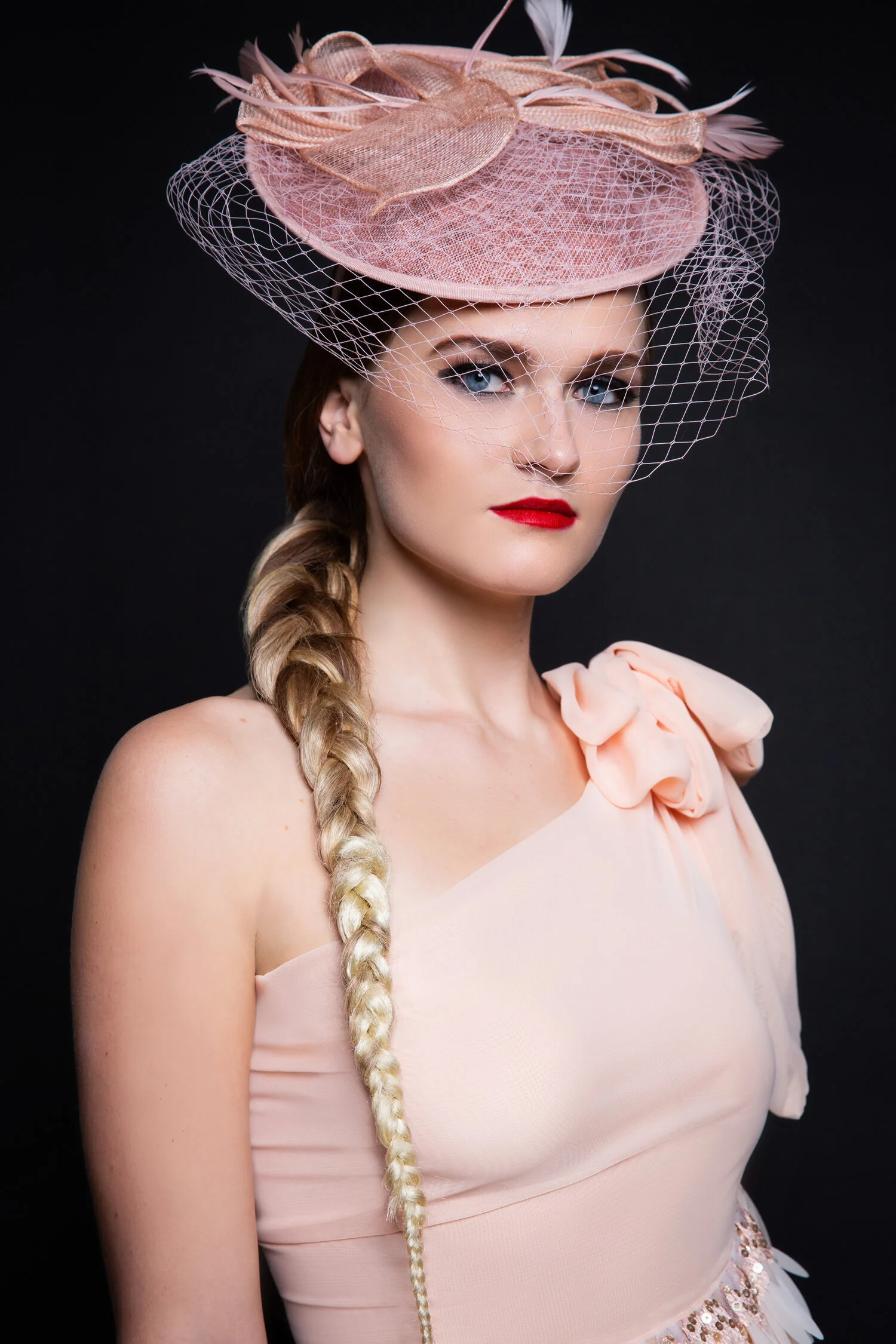 Woman wearing a pink fascinator with netting and a one-shoulder dress, styled with a side braid and red lipstick.