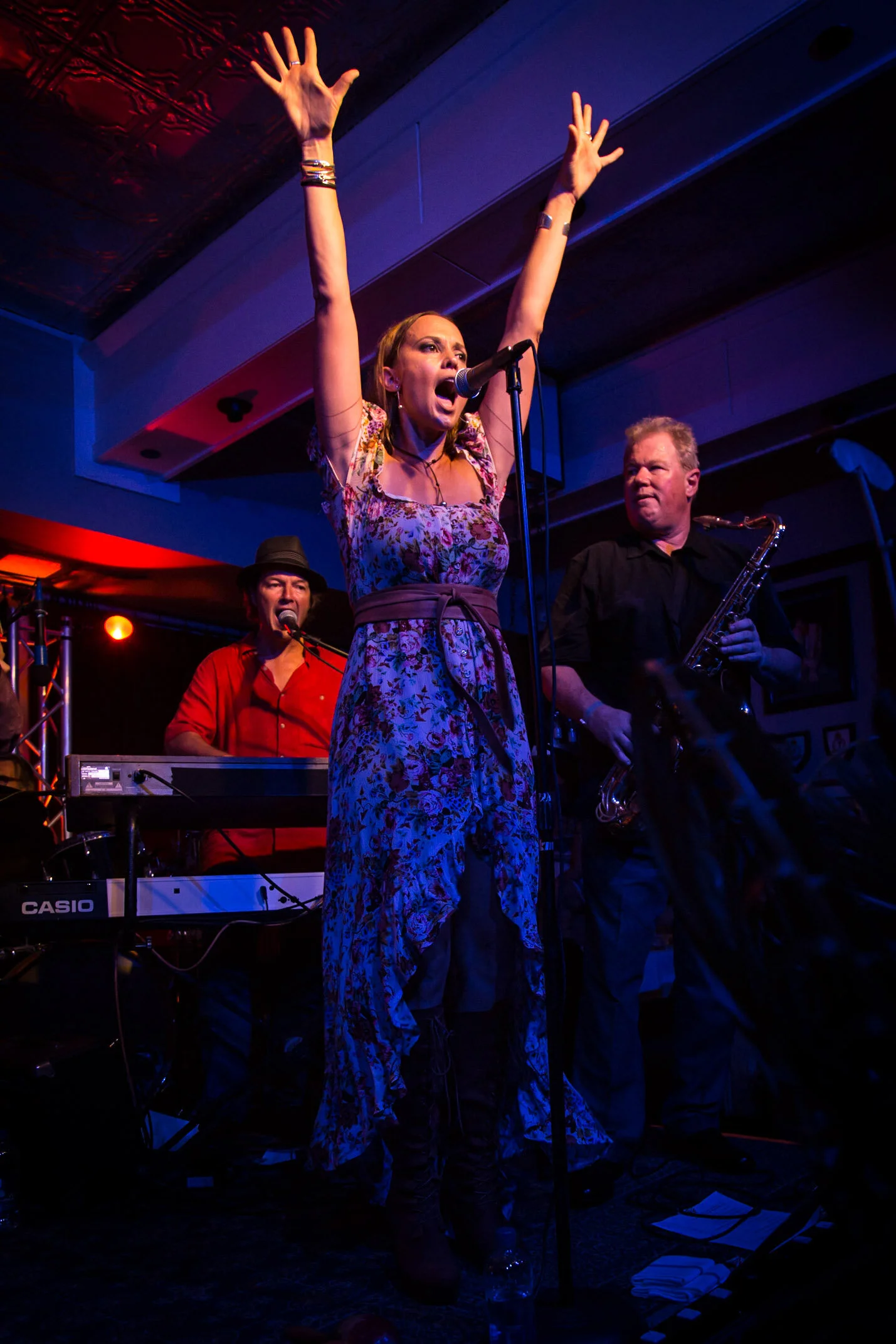 A woman in a floral dress singing passionately on stage with a microphone, surrounded by a keyboard player and a saxophonist, under colorful stage lighting.