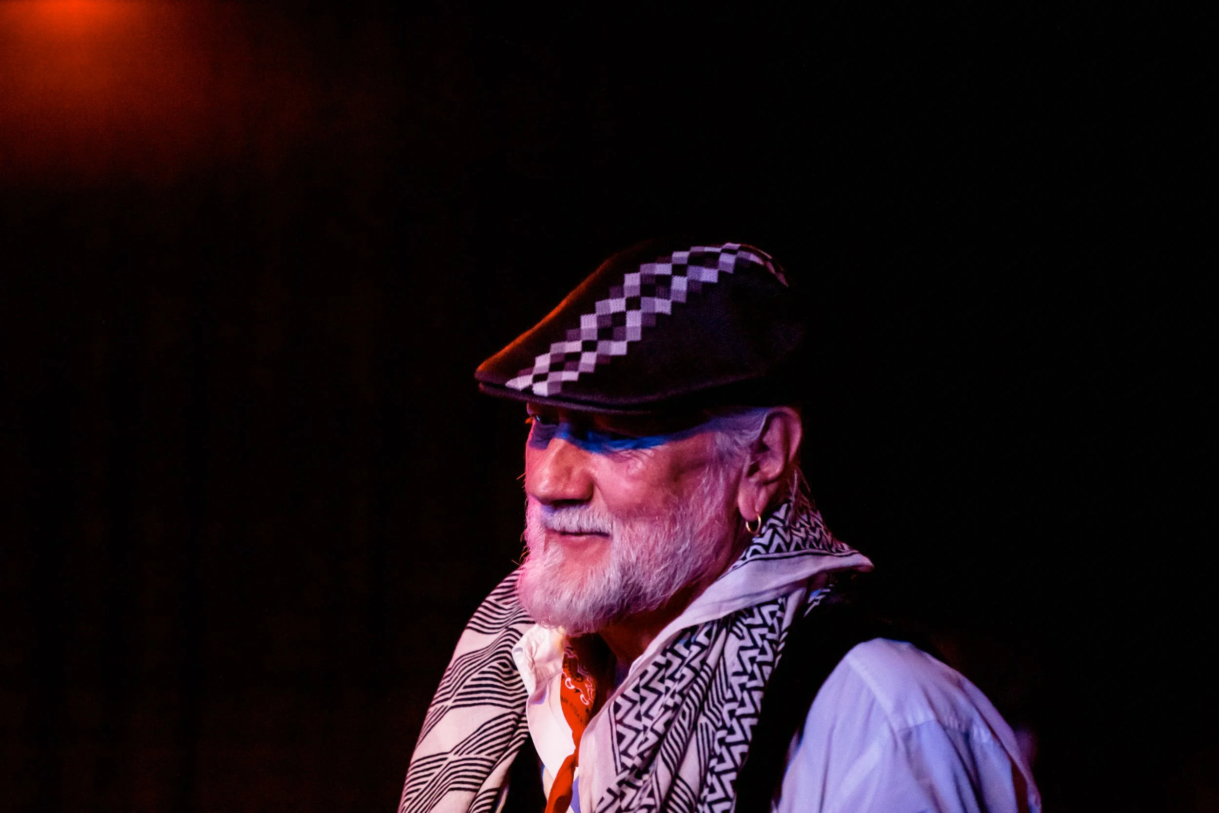 Bearded man wearing a patterned flat cap, white shirt, and black and white scarf at night.