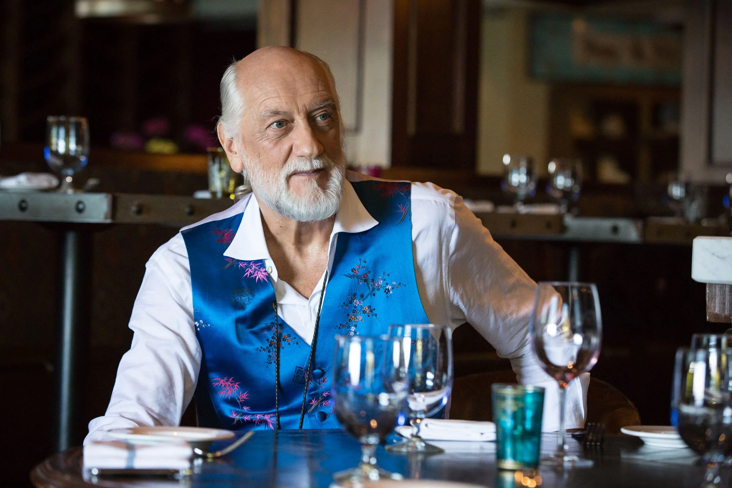 A man with a white beard wearing a bright blue vest sits at a table in a restaurant, surrounded by empty glasses and plates.