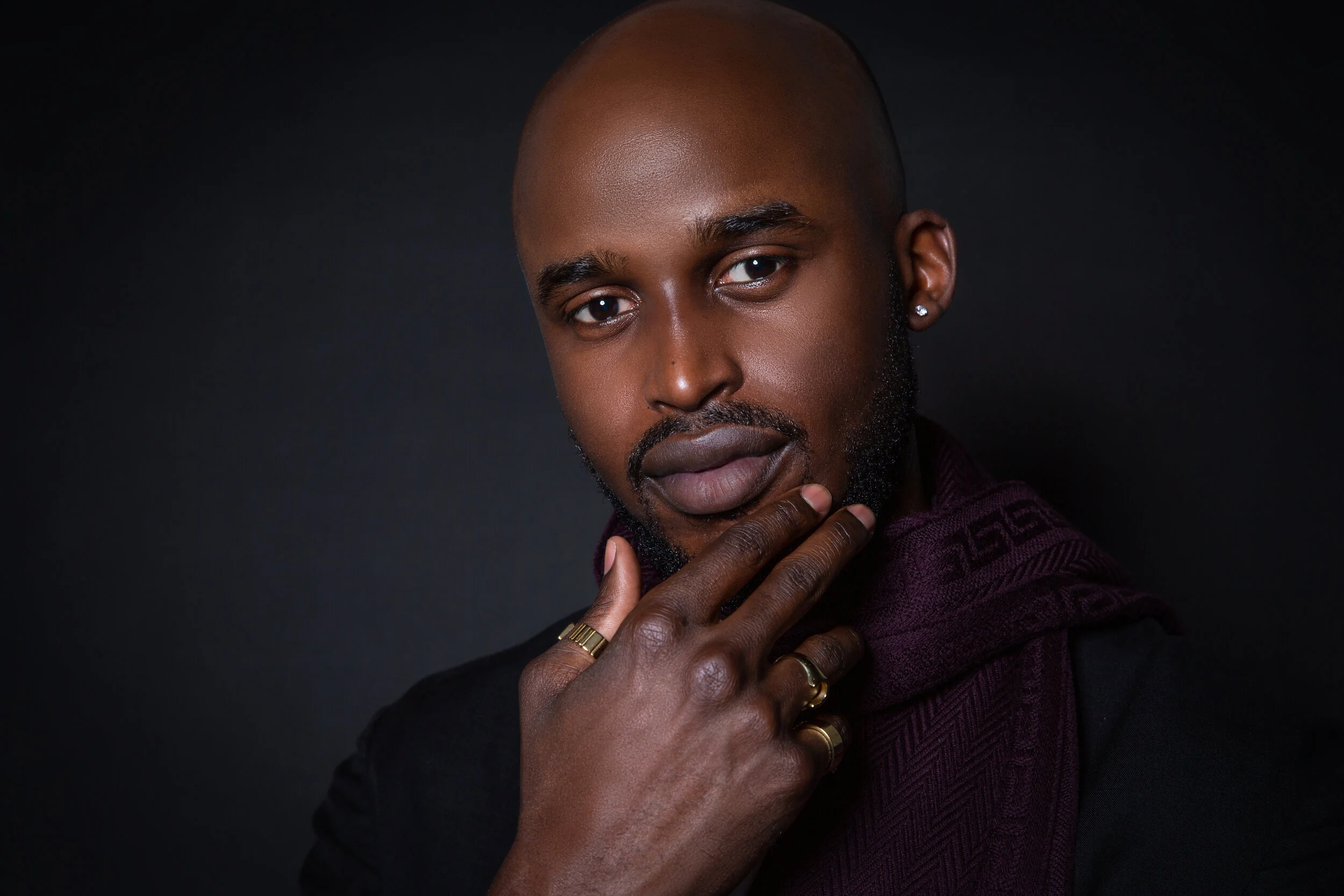 Man with a bald head and beard, wearing rings and purple scarf, with hand on chin, against a dark background.
