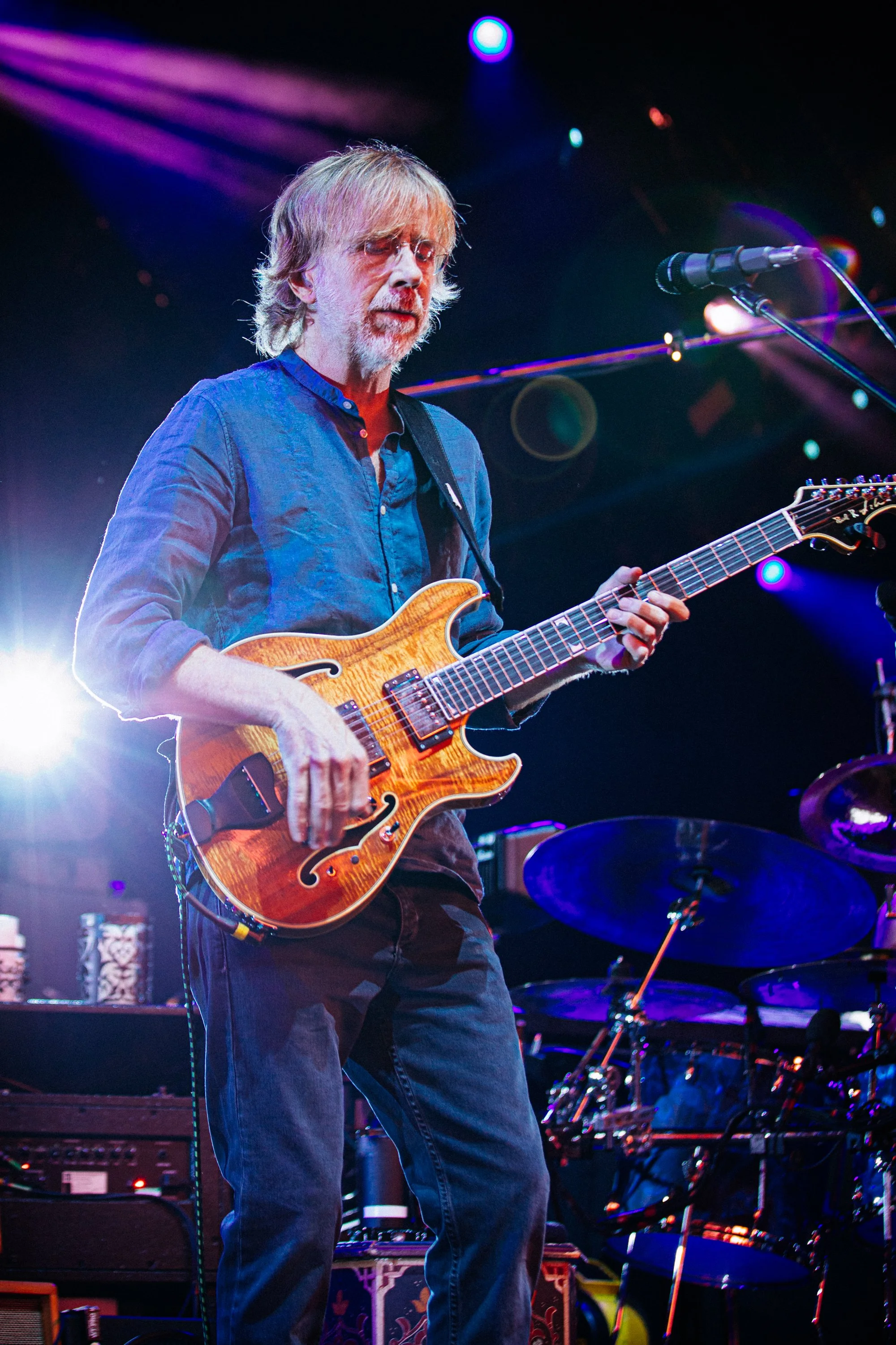 Guitarist performing on stage with electric guitar, stage lights, and drums in background.