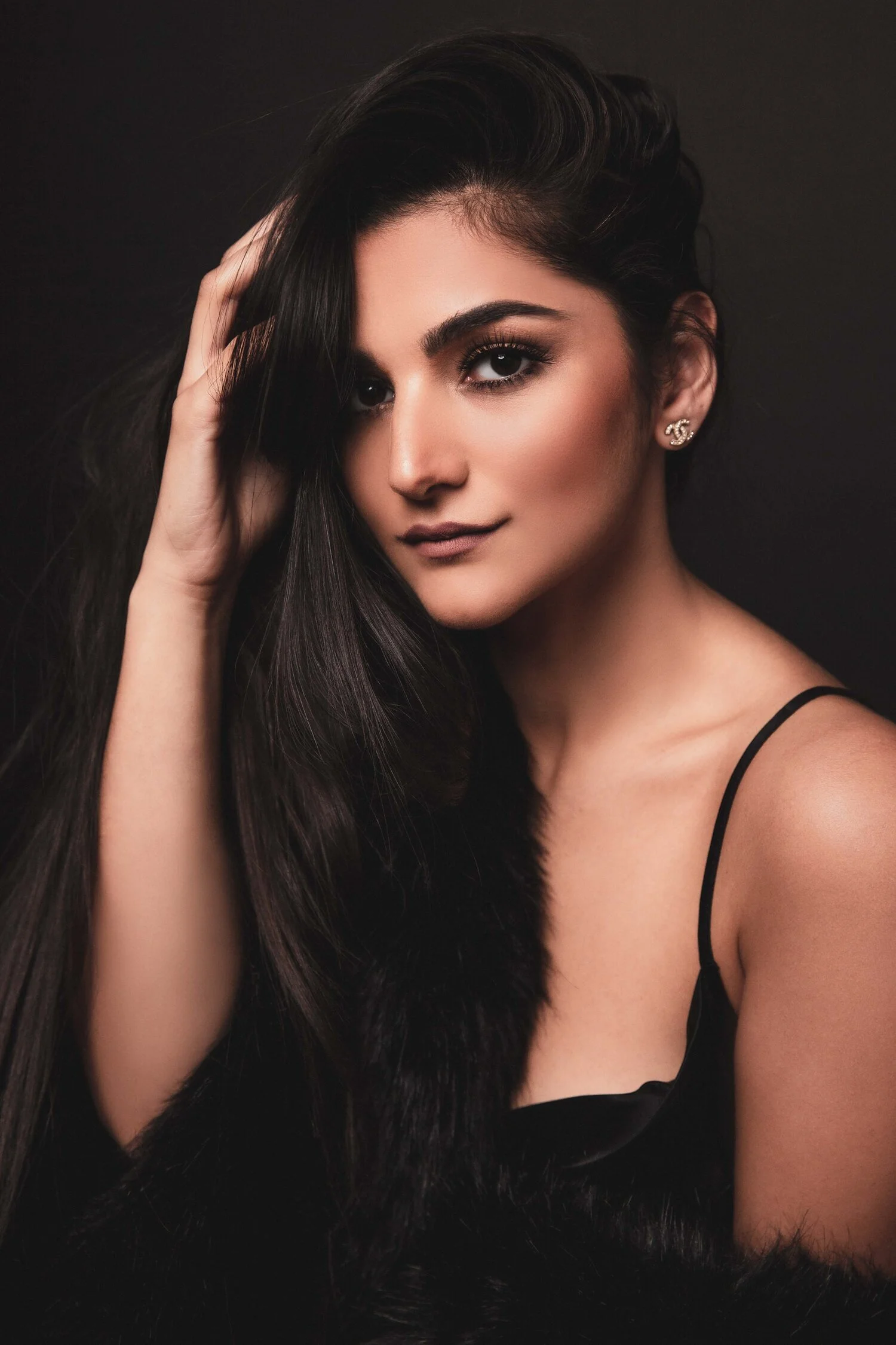 Portrait of a woman with long black hair, wearing a black outfit and earrings against a dark background, looking confidently at the camera.