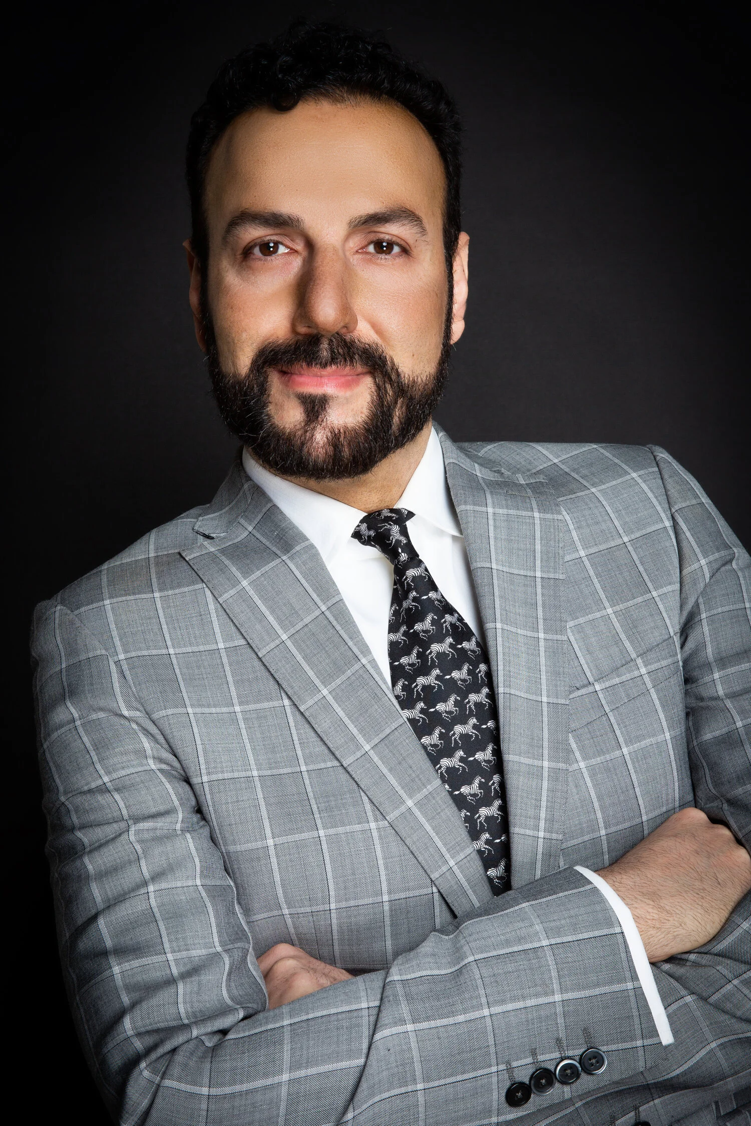Man in a gray checkered suit with a patterned tie, arms crossed, on a dark background.