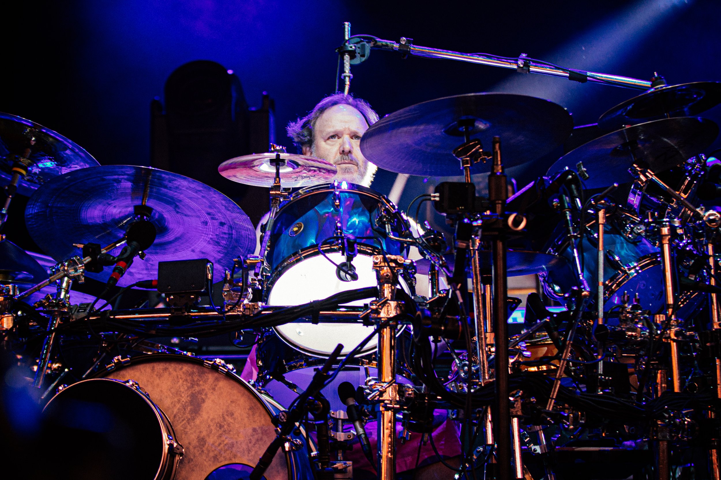 Drummer playing a drum kit with cymbals and microphones on stage under blue and purple lighting.