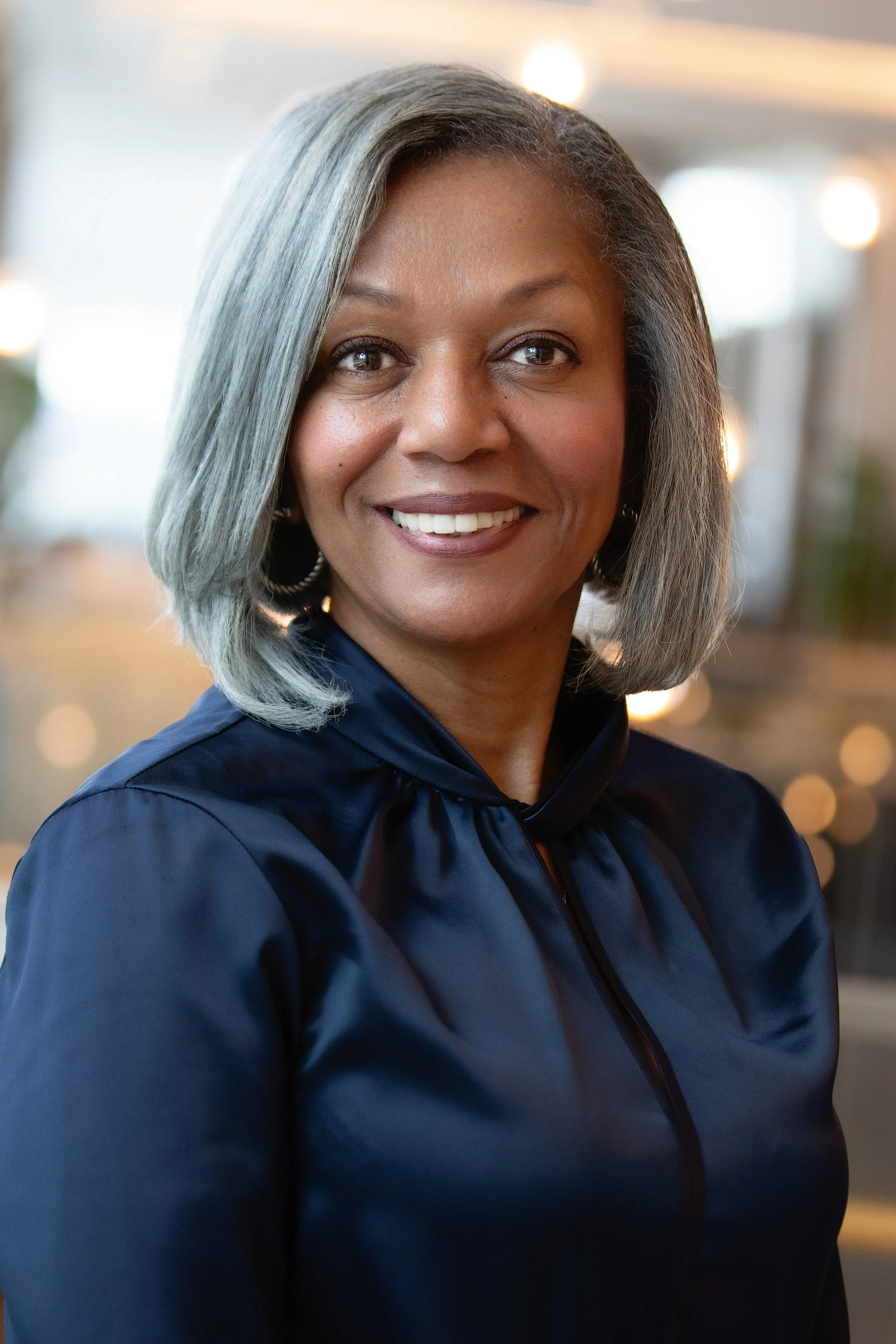 Portrait of a woman with gray hair smiling, wearing a navy blue top, in front of a blurred indoor background with soft lighting.