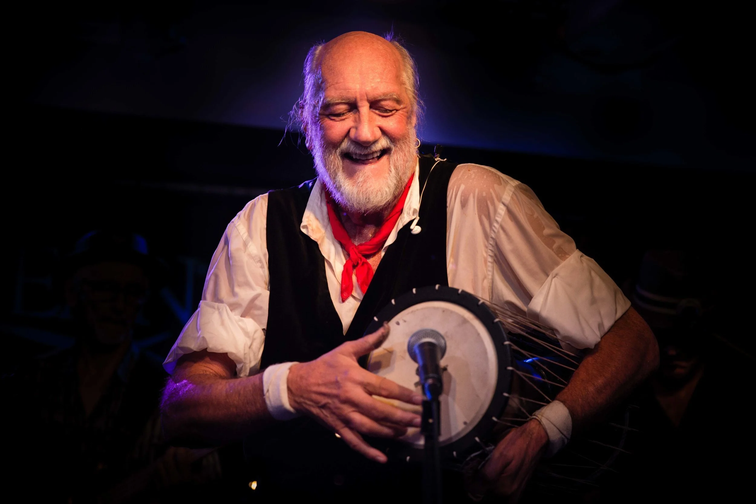 Person playing a drum on stage, wearing a vest and red scarf, with a microphone.