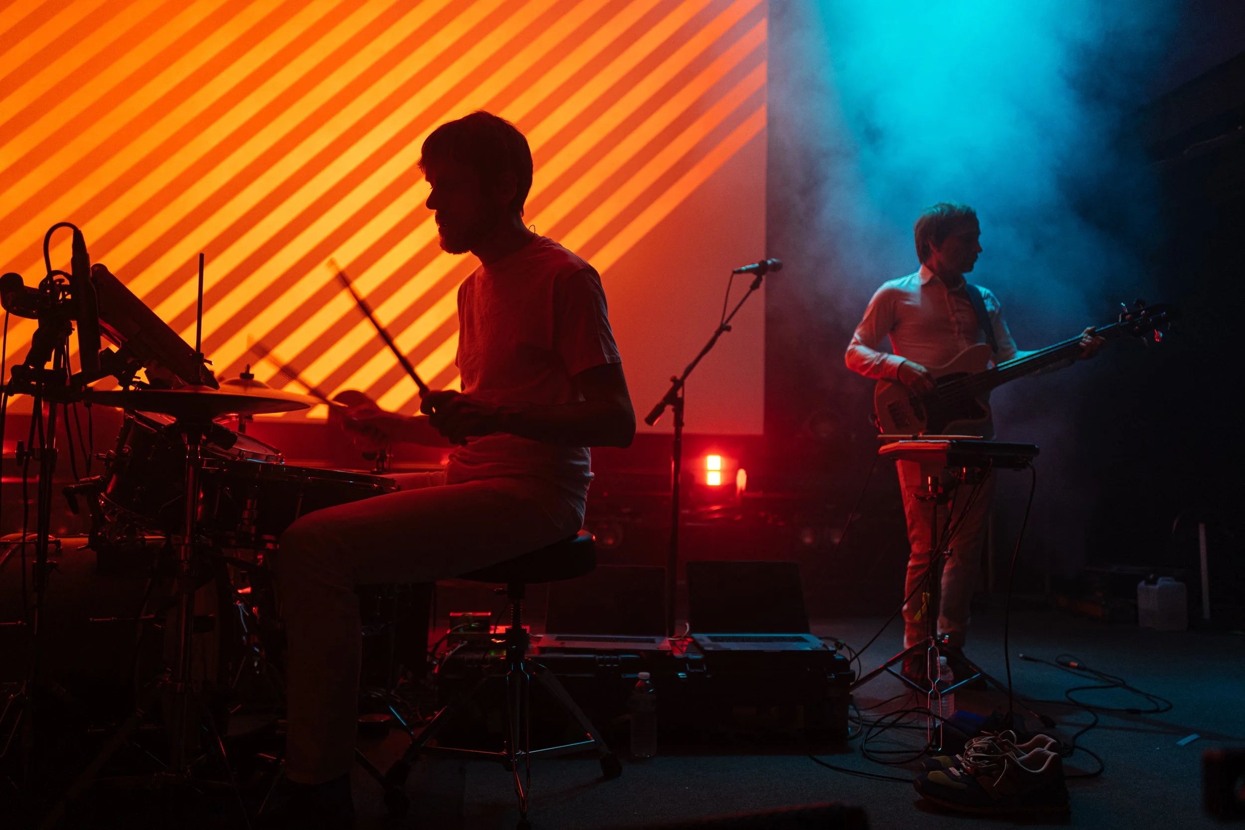 Musicians performing on stage with colorful lighting, one playing drums and the other playing bass guitar.