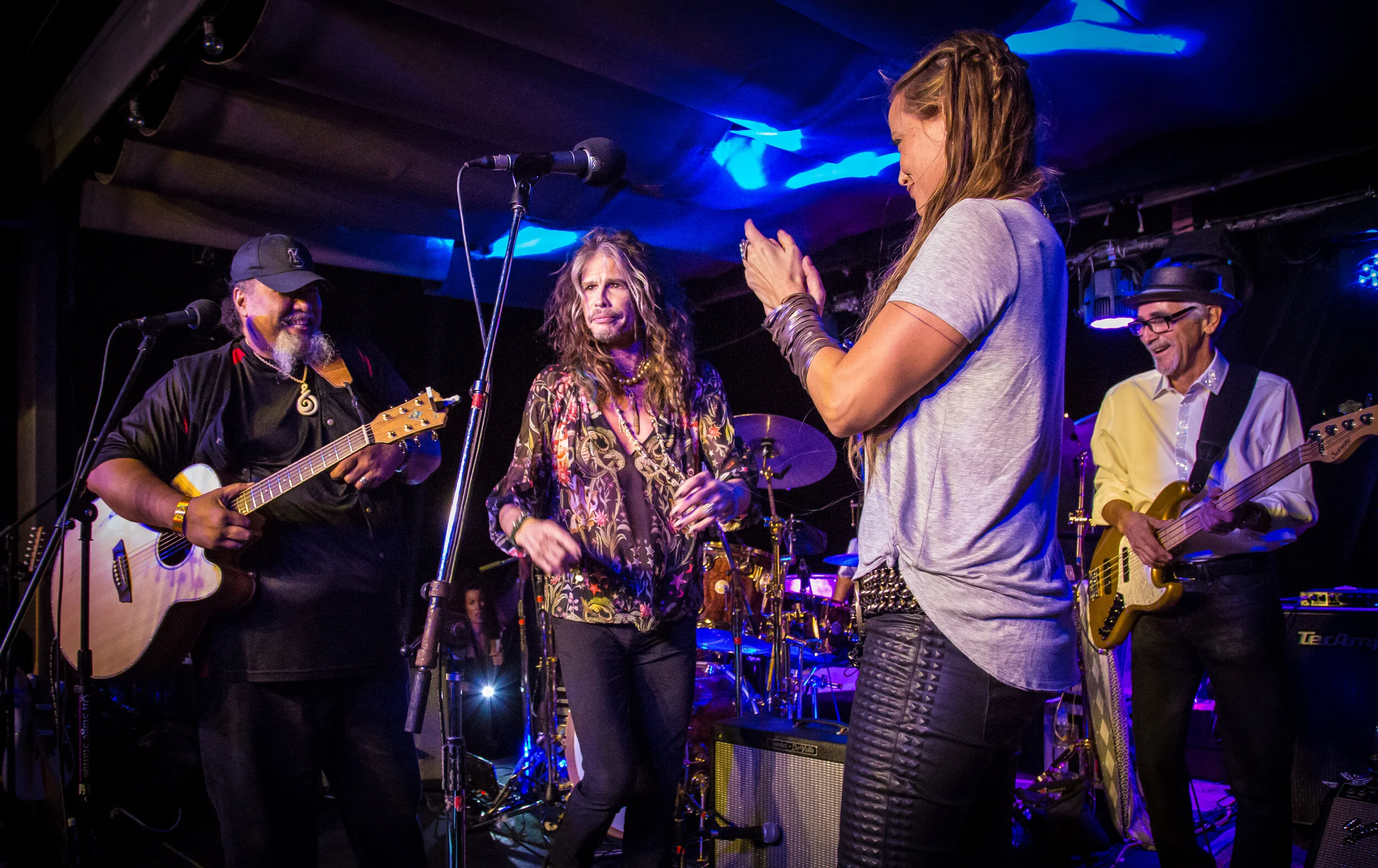 Musicians on stage, including a guitarist, a singer, and a woman clapping. The guitar player is wearing a black shirt and cap, the singer has long hair and a patterned shirt, and the woman in a gray shirt is clapping. Stage lights illuminate the scen
