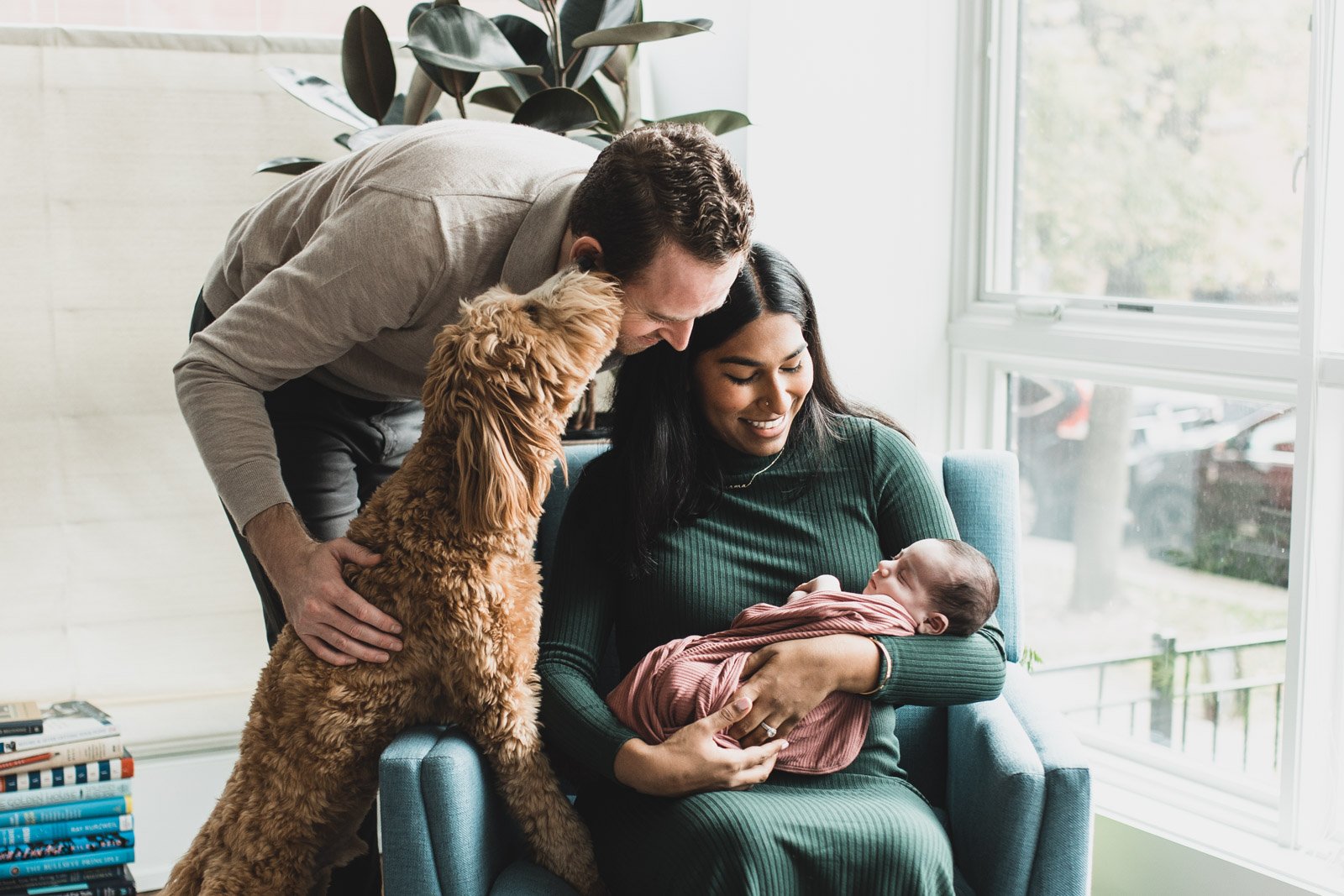 mom and dad looking down at newborn baby with dog licking dads face