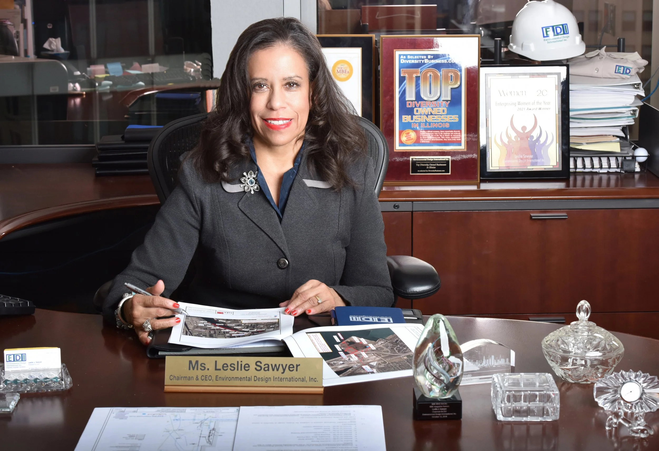 A woman sitting at a large wooden desk in an office, holding a pen and looking at the camera. She has dark brown hair, is wearing a gray blazer with a brooch on the lapel, and has a nameplate reading Ms. Leslie Sawyer. The desk has various papers, a glass paperweight, a decorative glass object, and awards and framed certificates in the background.