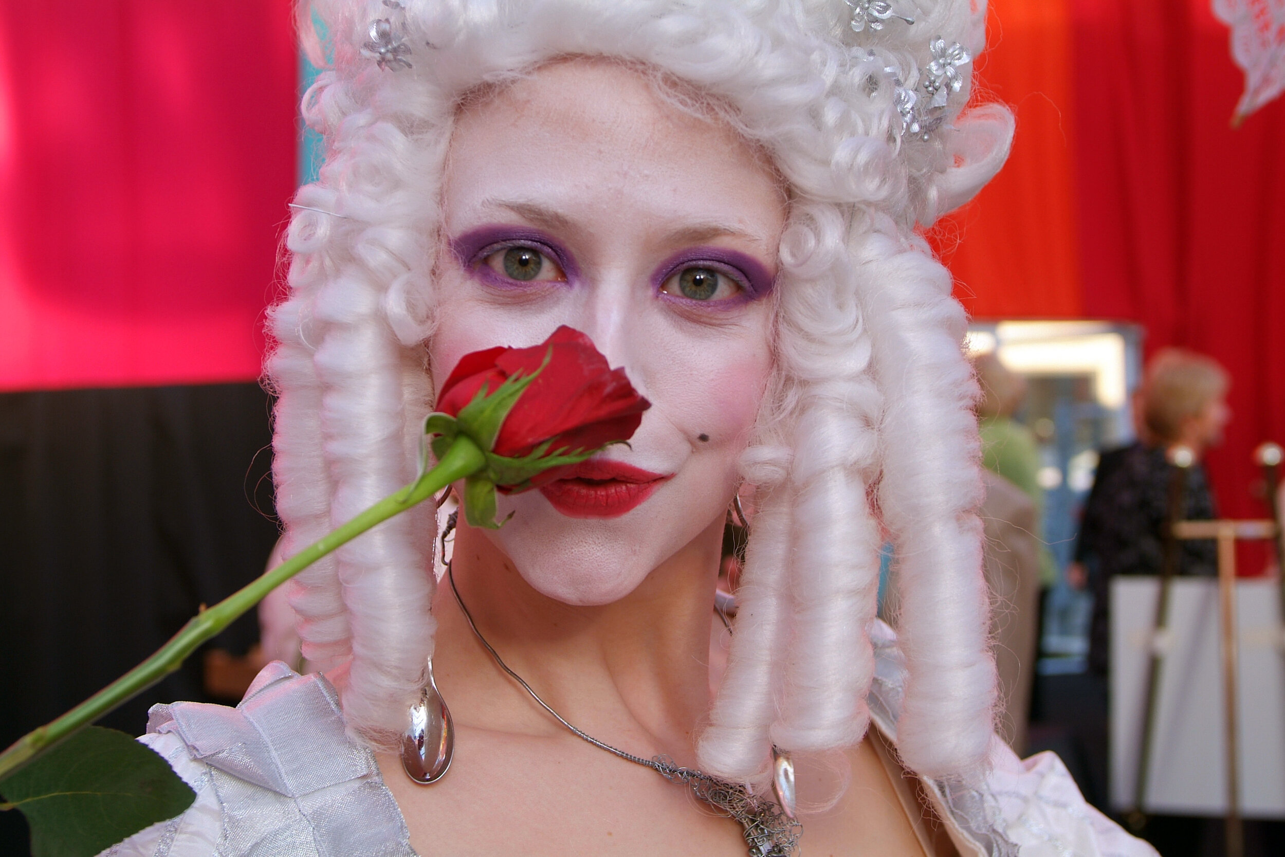 Person dressed as a character with a white curly wig, purple eye makeup, and red lipstick, holds a red rose near their face, with an ornate necklace visible, in a setting with red curtains and blurred people in the background.