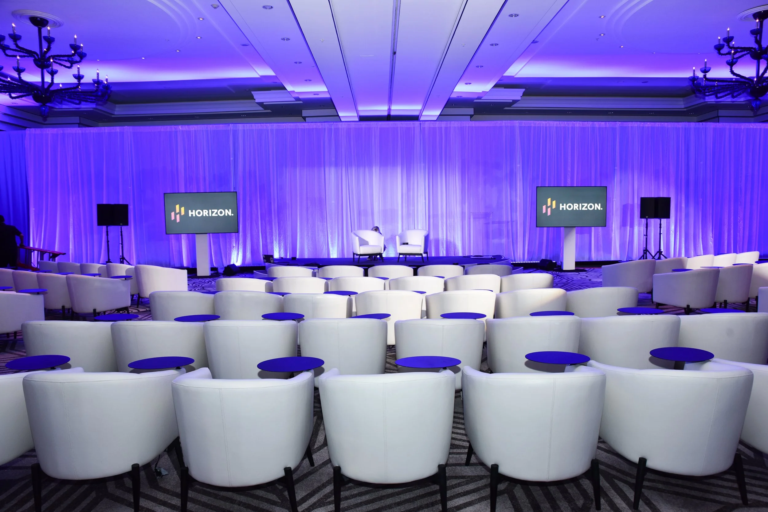 Empty conference room with white chairs, small blue tables, a stage with two white chairs, and two screens displaying 'HORIZON' logo, illuminated in purple lighting.