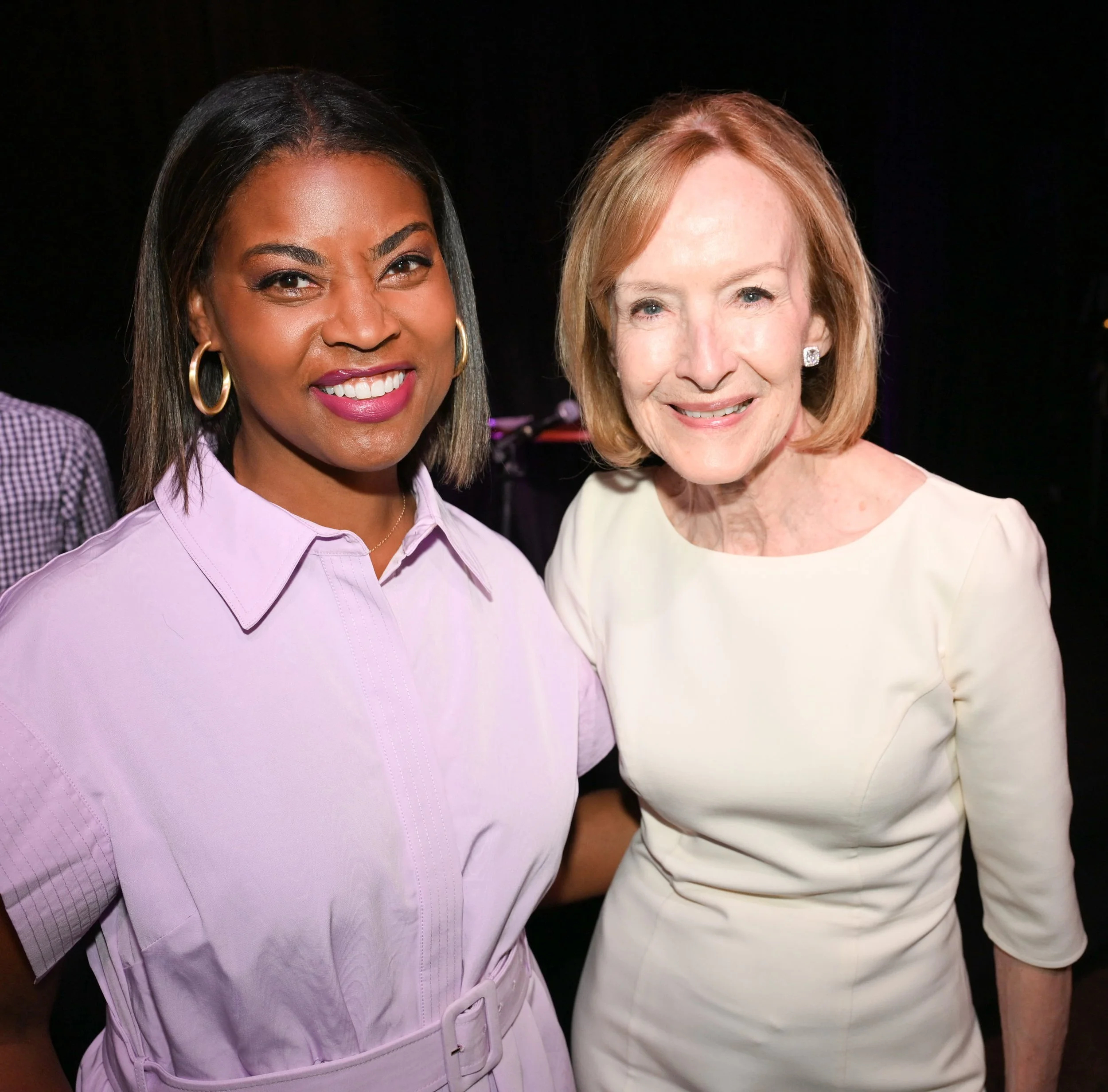 Two women smiling, standing close together at an indoor event, one wearing a light purple dress and the other in a light-colored dress.