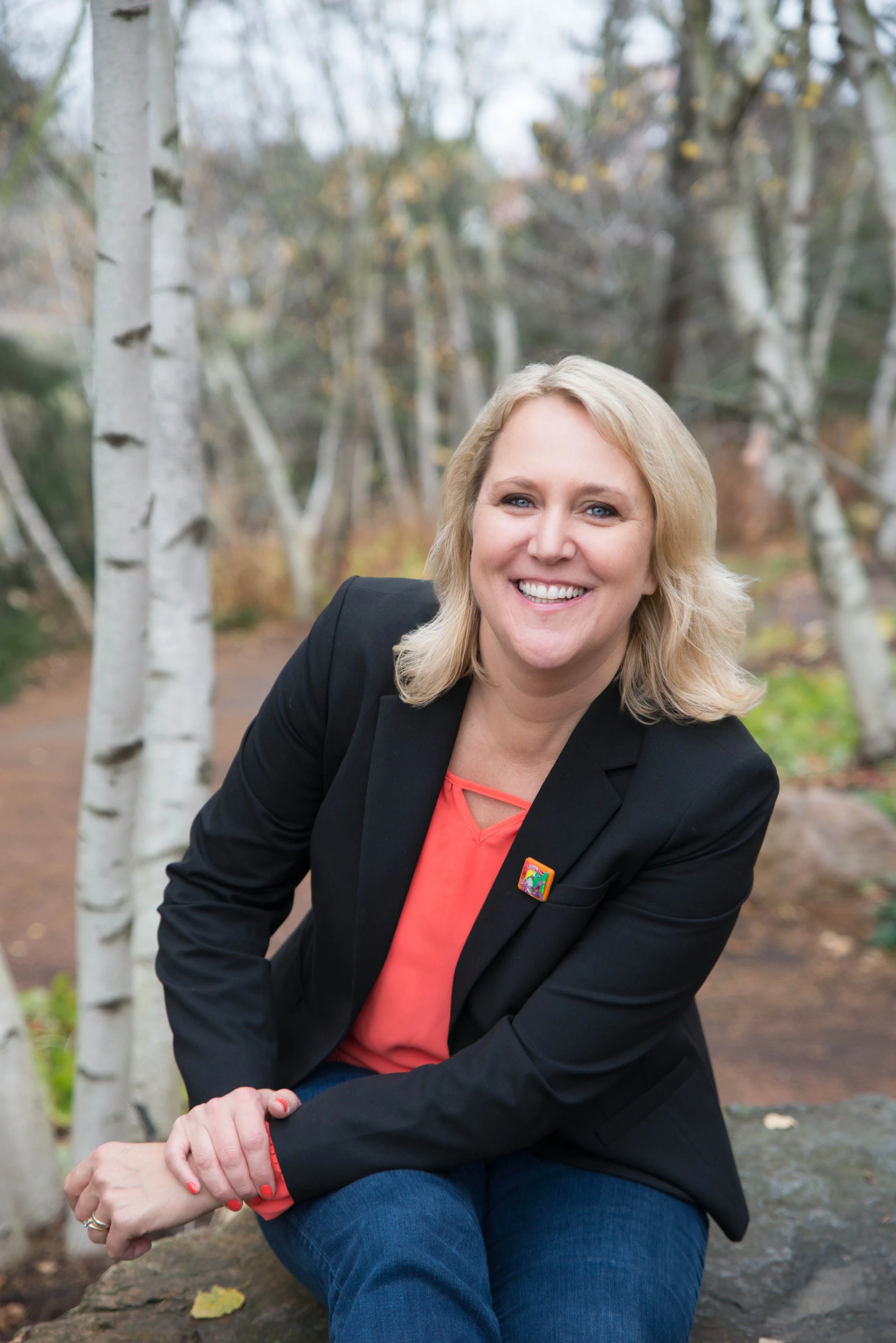 A woman with shoulder-length blonde hair, wearing a black blazer with a rainbow badge, and a coral blouse, sitting outdoors on a rock in a wooded area with white trees and autumn leaves.