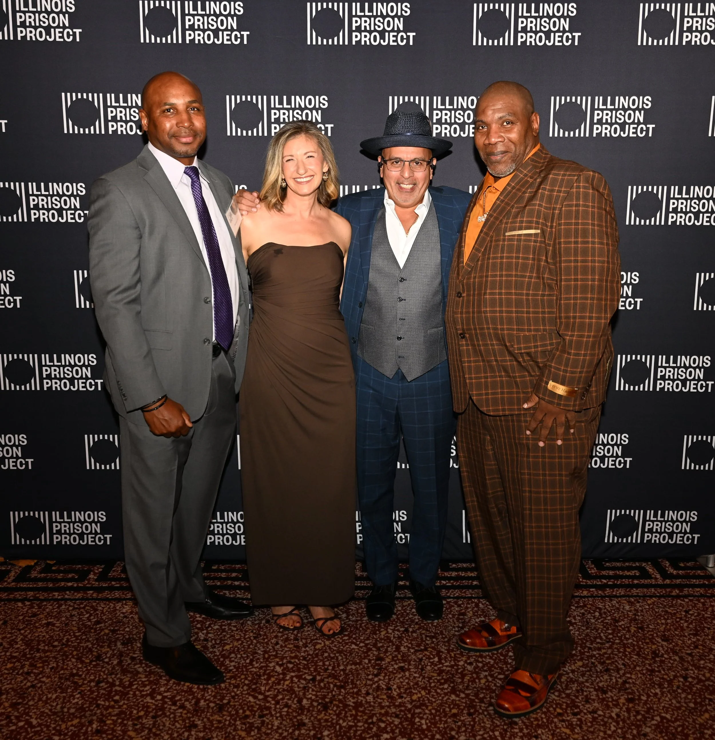 Four people posing together in front of a backdrop with the Illinois Prison Project logo. They are dressed in formal attire, smiling, and standing close together.