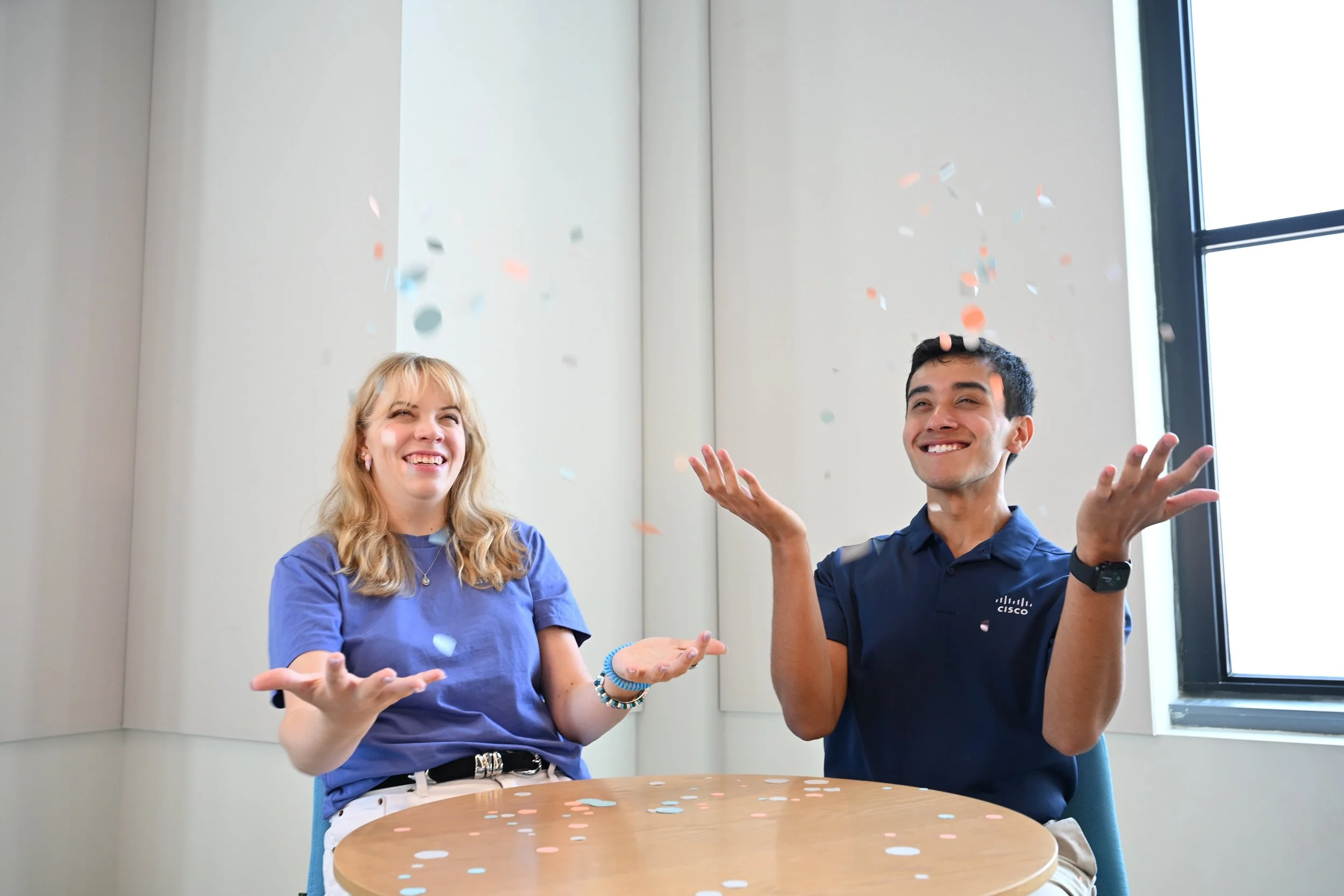 Two young adults, a woman and a man, sitting at a round table, celebrating with confetti falling around them, smiling and looking joyful.