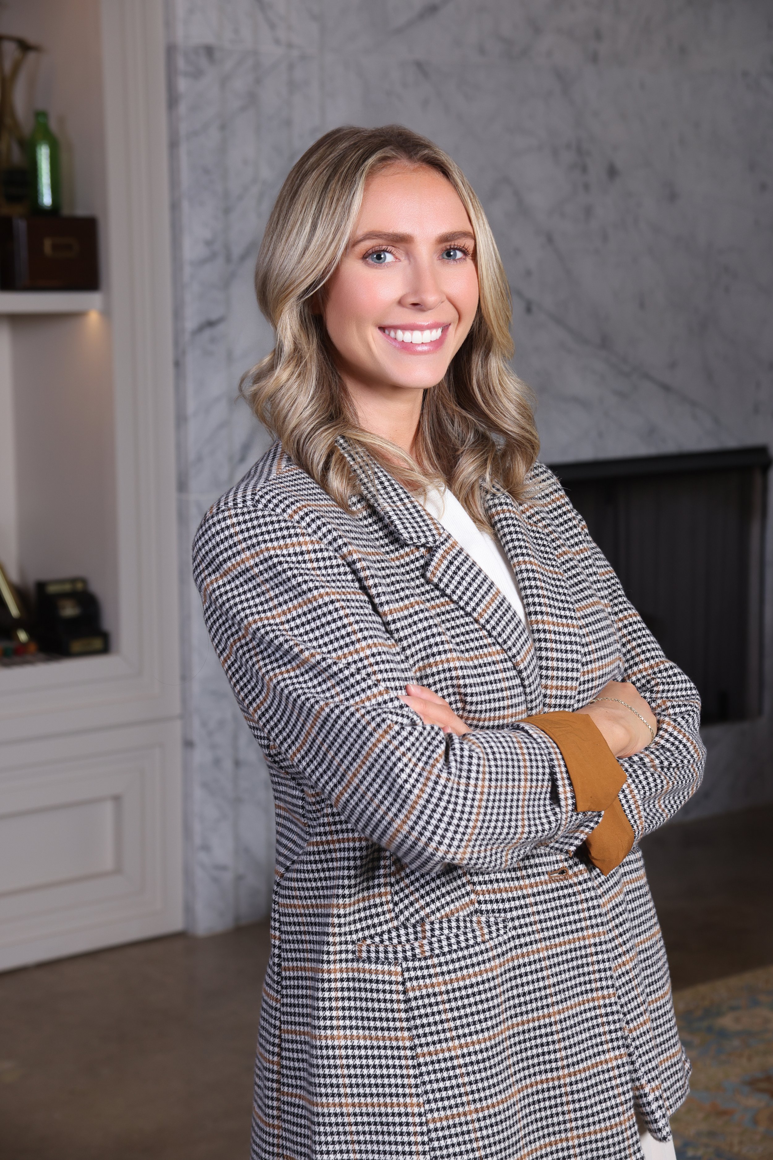 A woman with blonde hair wearing a checked blazer and white top, smiling with arms crossed, standing indoors in front of a marble wall and fireplace.
