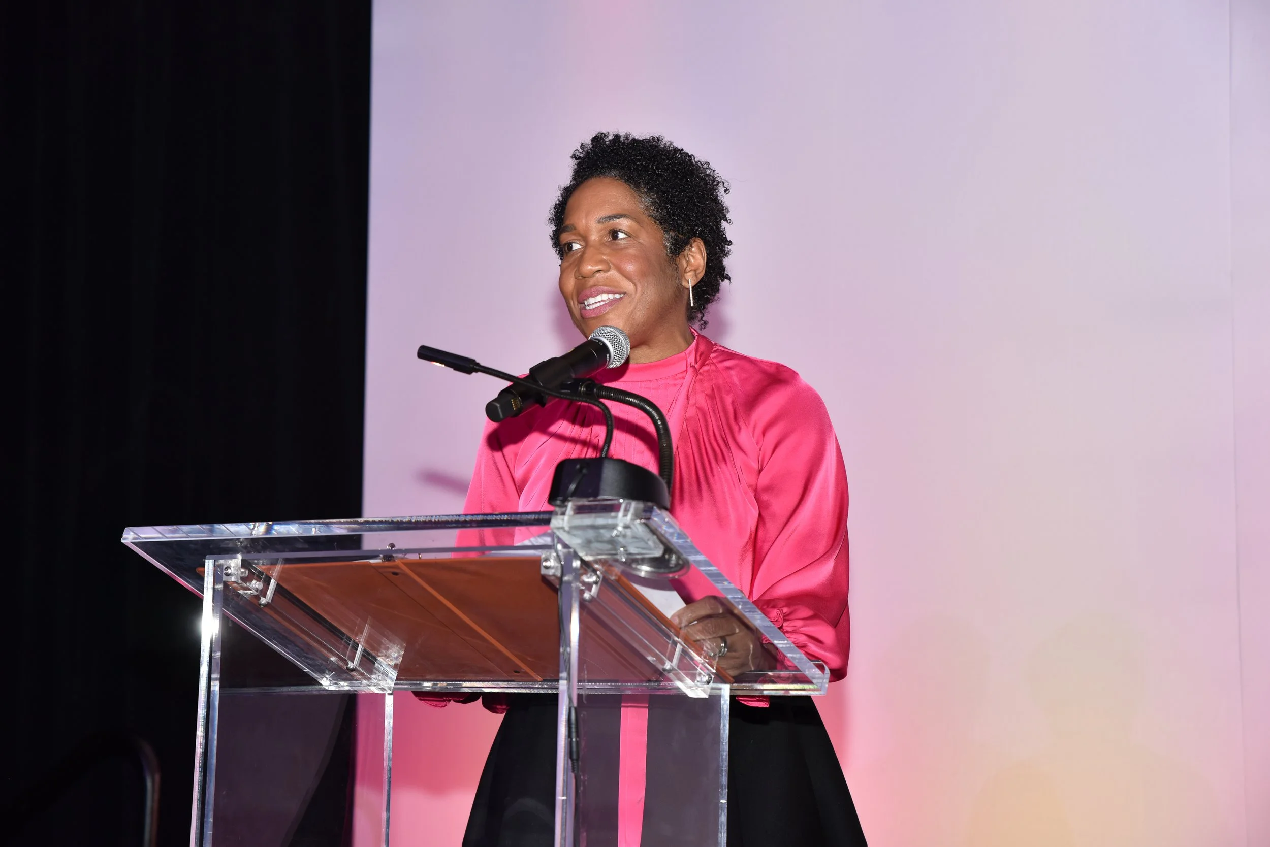 A woman with short, curly black hair wearing a pink satin blouse speaking at a clear acrylic podium with a microphone, against a plain light purple background.