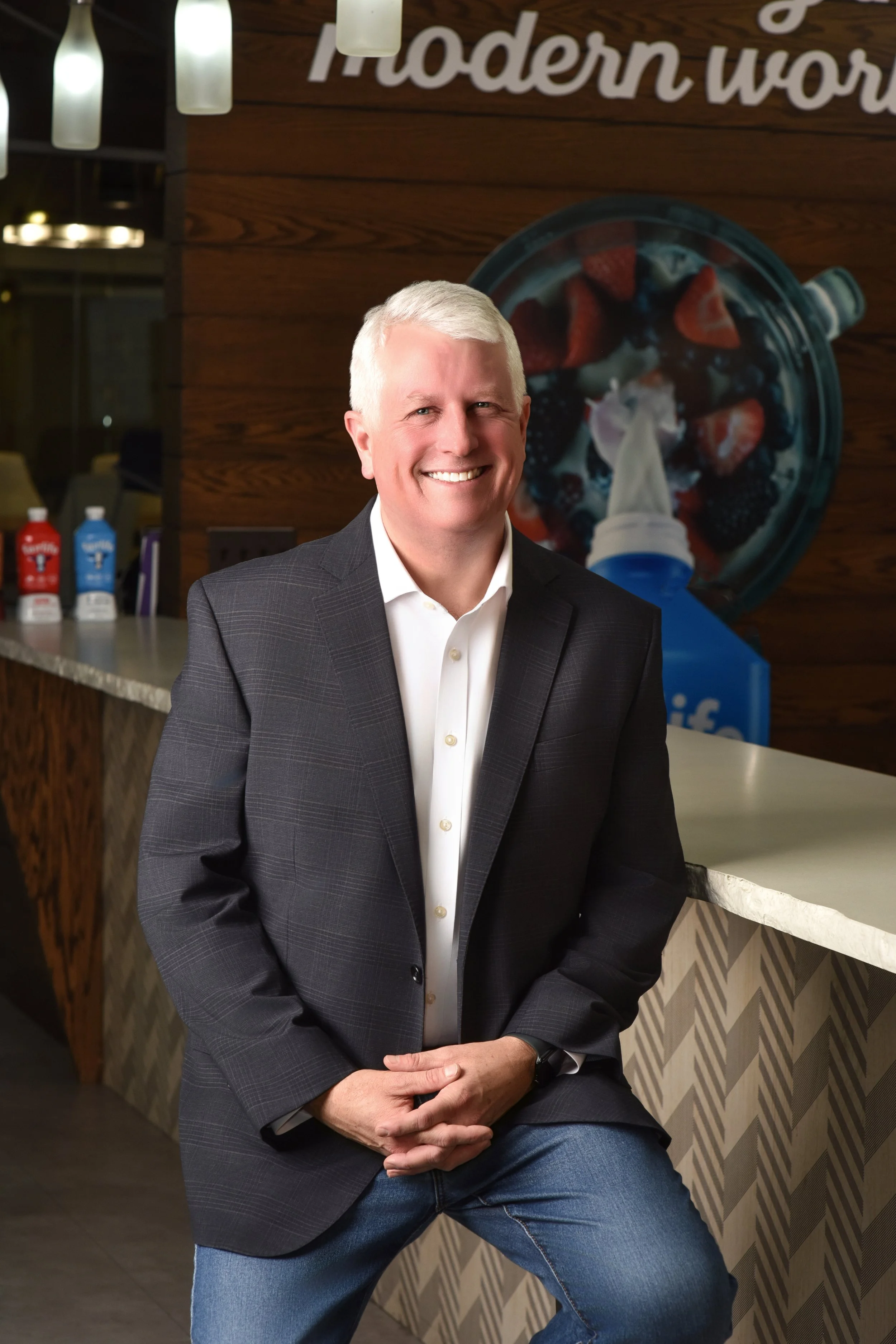 A smiling man with short gray hair wearing a white button-up shirt and a dark blazer, leaning against a bar counter in a modern setting, with a large colorful image of a fruit dessert and a bottle of yogurt in the background.