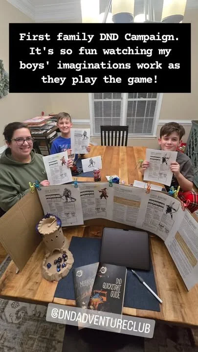 A family sitting at a dining table with rolled-up D&D campaign sheets attached by clips. The mother and two boys are smiling and holding dice and character sheets. There is a black digital tablet, two D&D guidebooks, and a decorative wooden dice hold