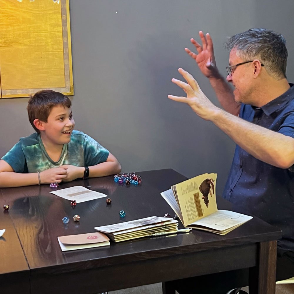A young boy and an adult man are playing a tabletop role-playing game with dice and books, both smiling and laughing.