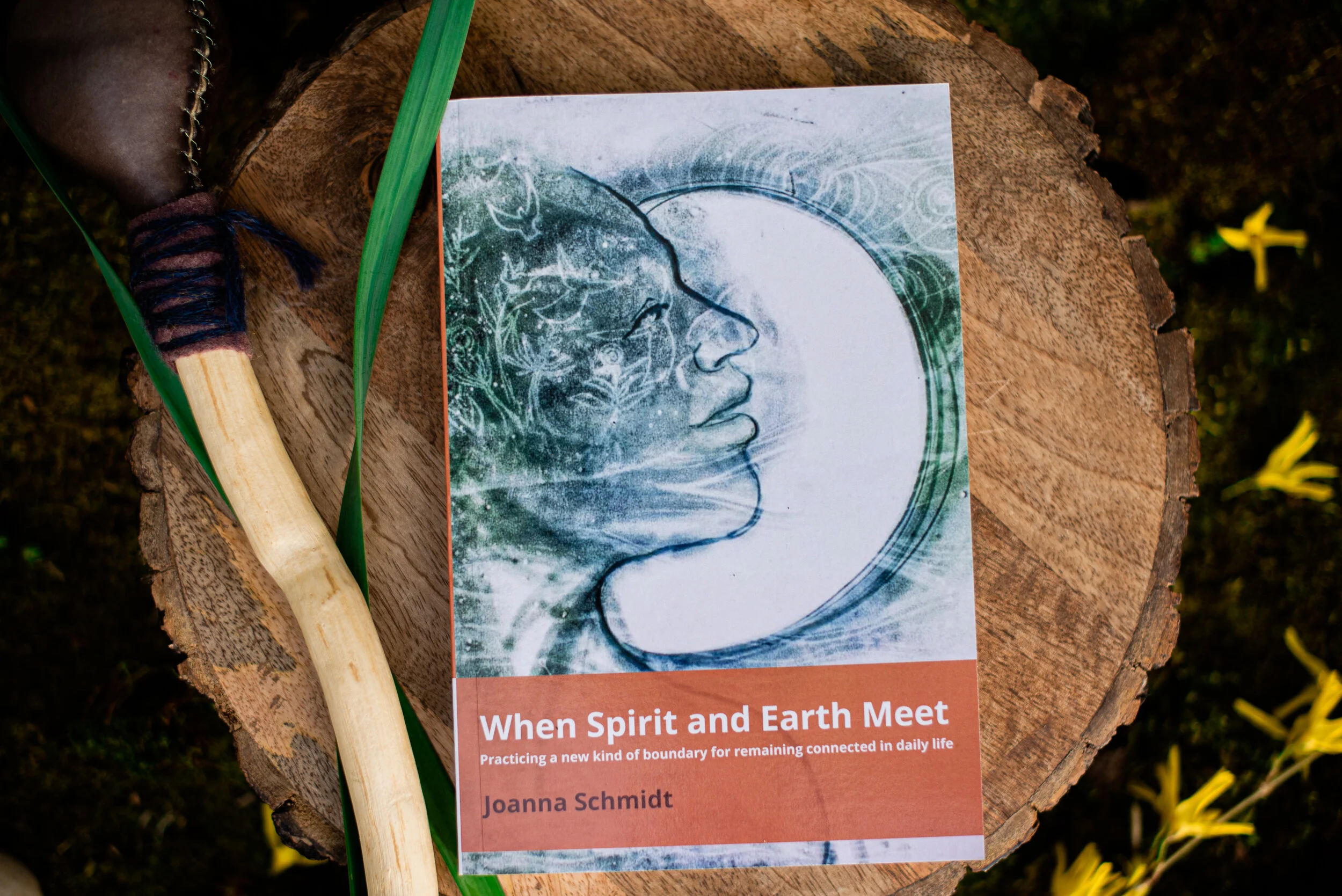 Paperback copy of "When Spirit and Earth Meet" by Joanna Schmidt on a natural wood table with a rattle next to it