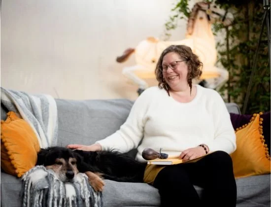 Animal Communicator, Joanna sitting on a grey sofa in a white sweater and black pants holding a journal and shamanic rattle. She is sitting with a black and tan dog who is laying comfortably by her side