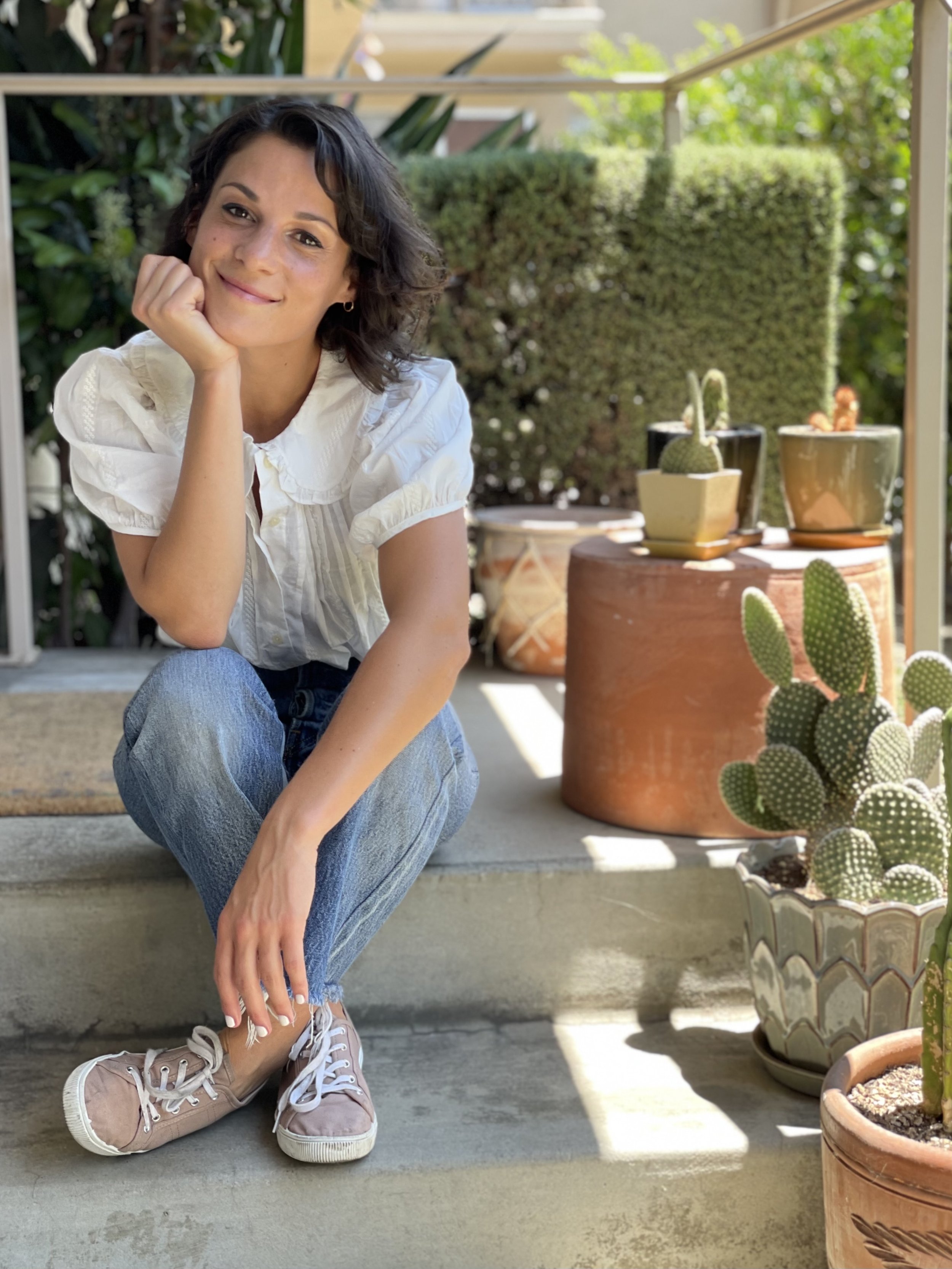 A woman with short dark hair, wearing a white blouse, jeans, and light pink sneakers, is sitting on a step outdoors, smiling and resting her chin on her hand. There are potted cacti and greenery in the background.