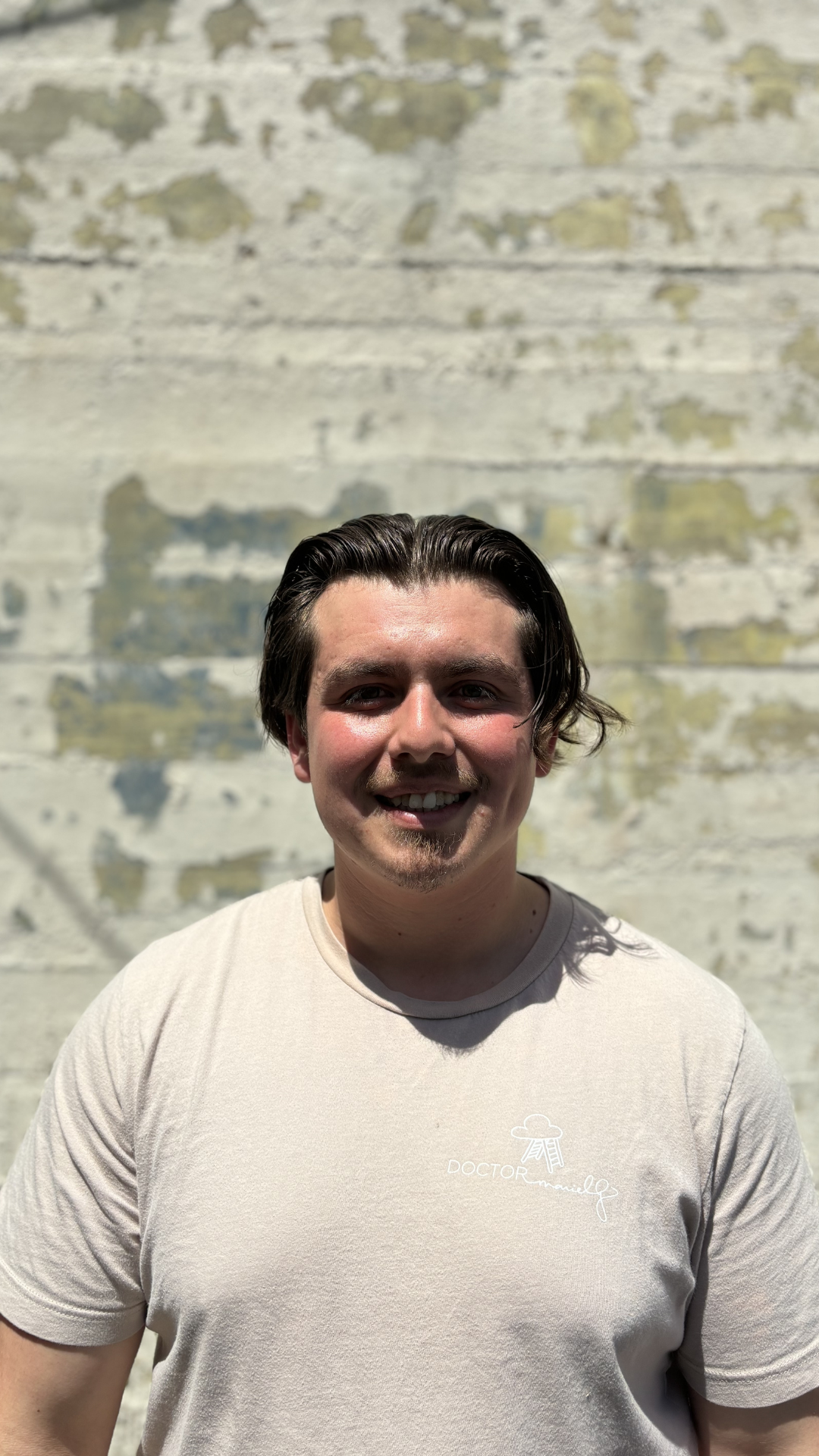 Smiling young man with dark hair standing outdoors in front of a weathered, off-white wall with peeling paint.