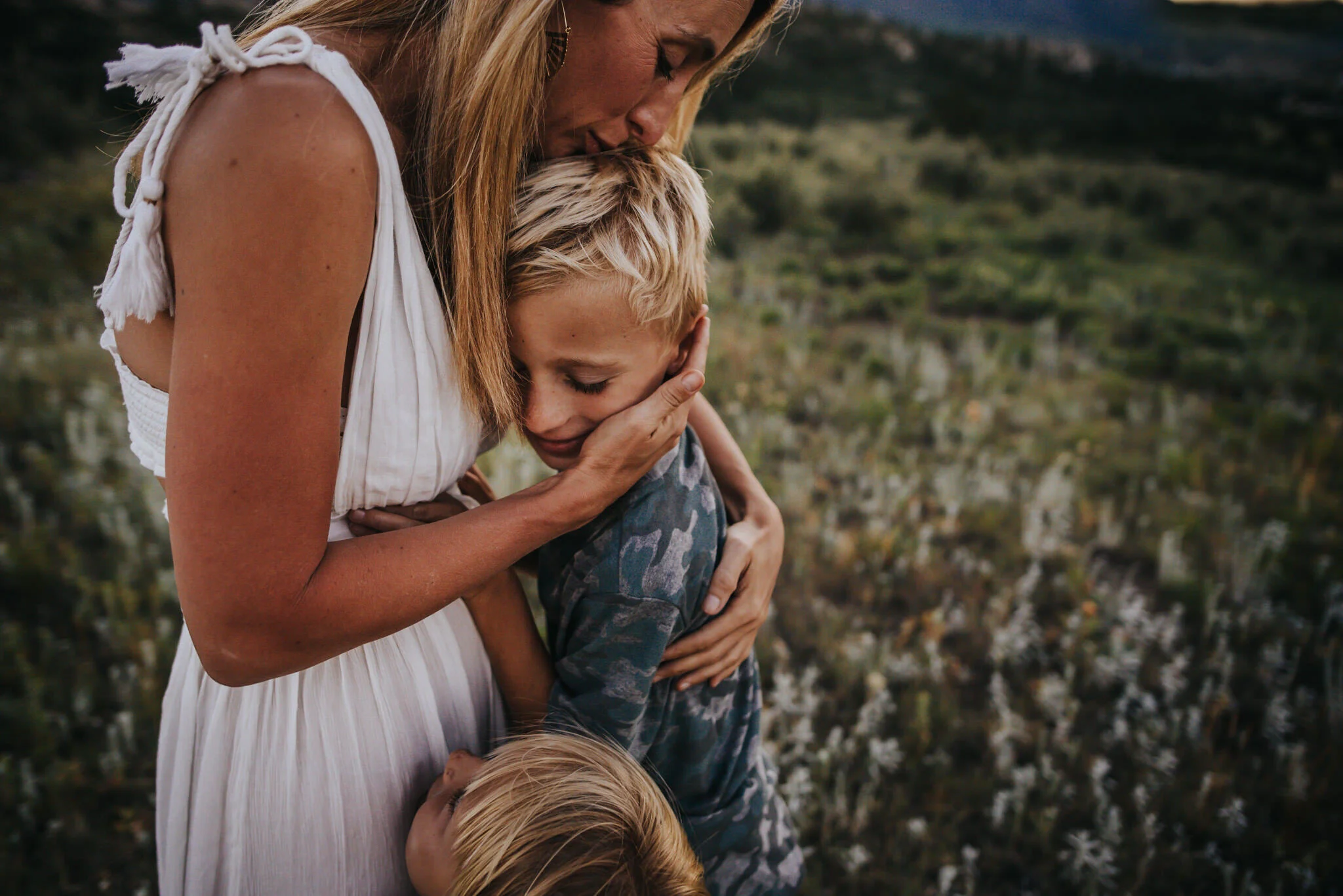 A mother holds her sons close to her and kisses one on the top of his head while the other looks up at her.