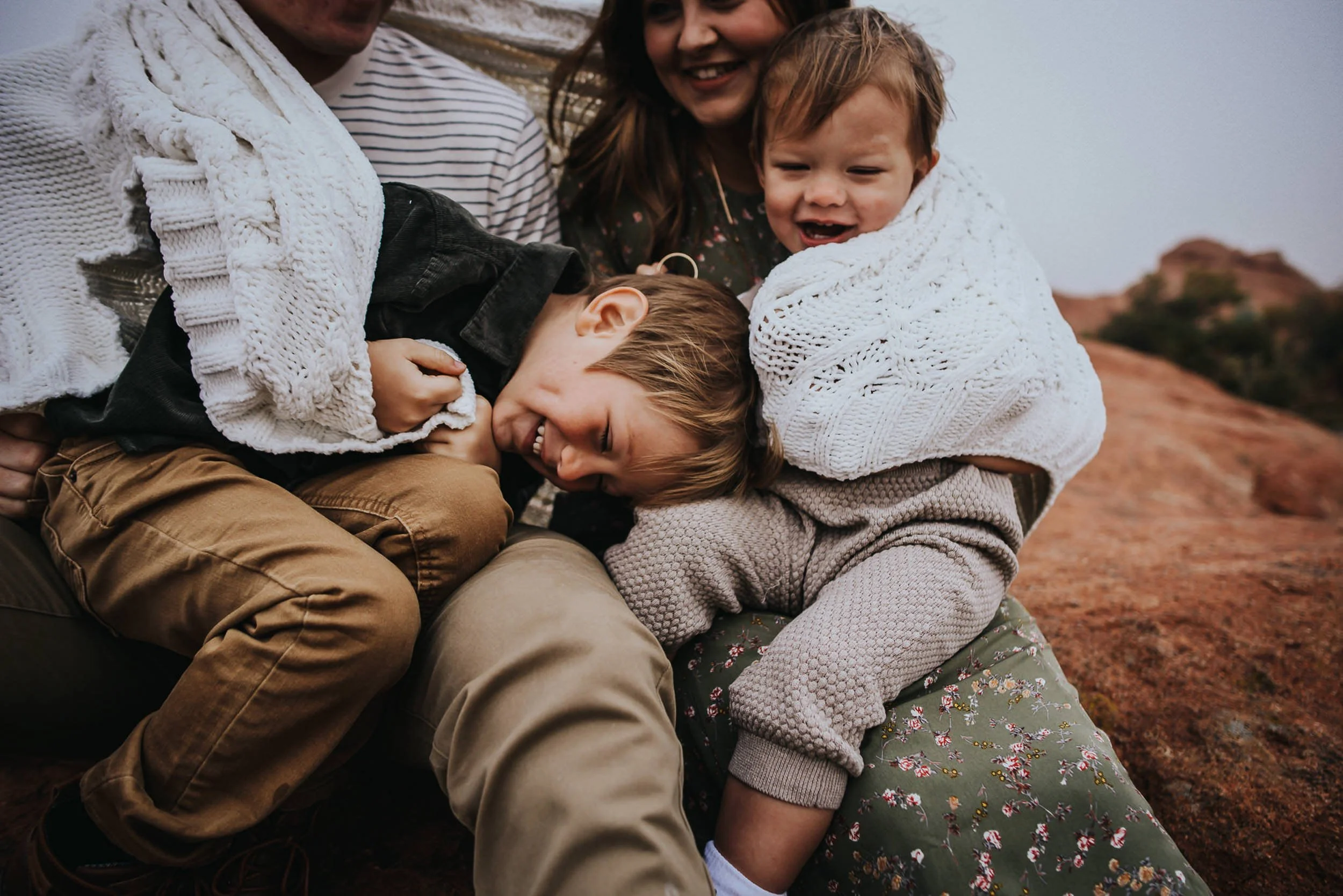 Three children piled together laughing during a family session, wearing coordinated earth tones in greens and neutrals.